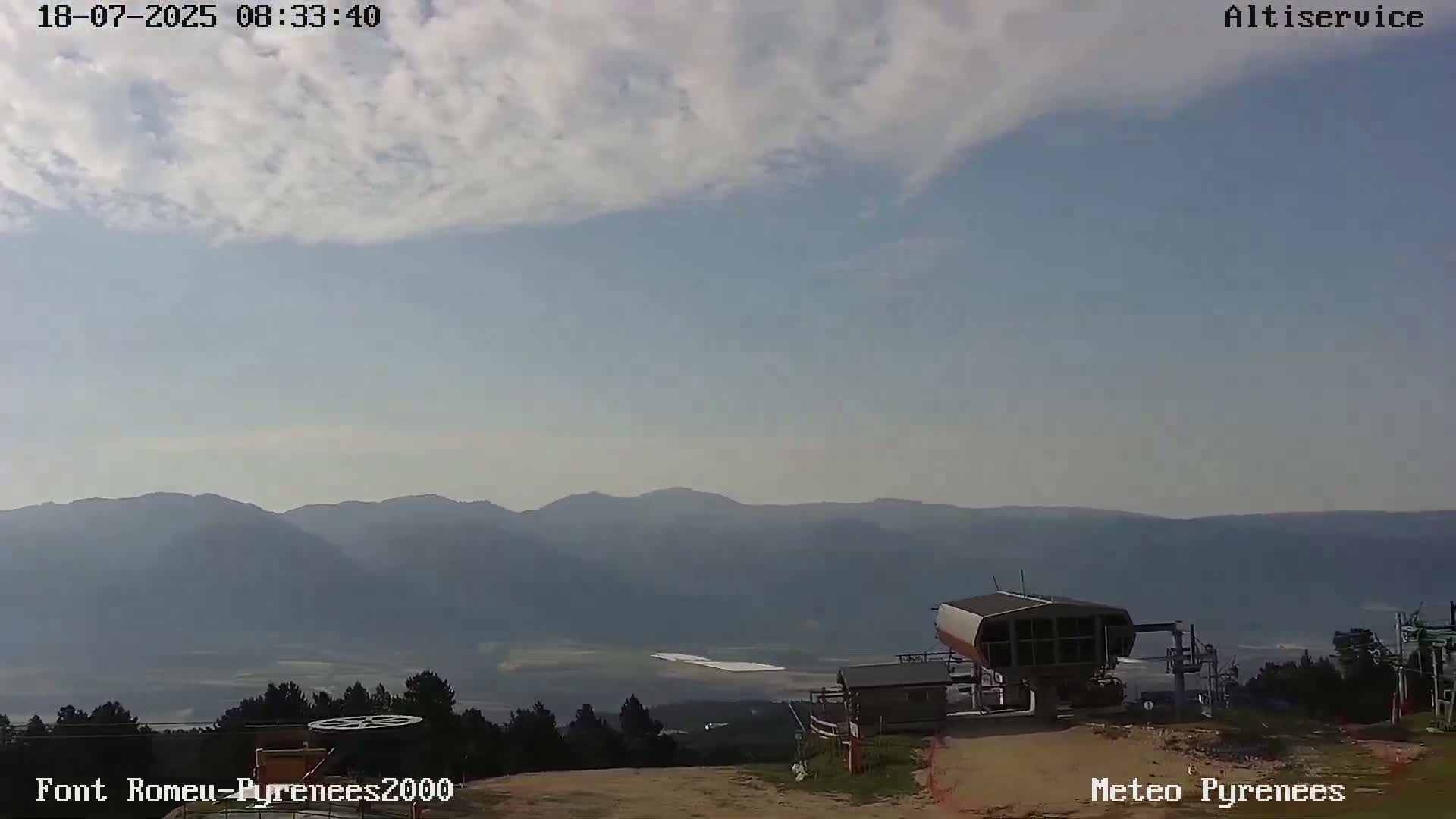A partly cloudy sky overlooks a mountain range and a ski lift in a hazy, mountainous landscape.