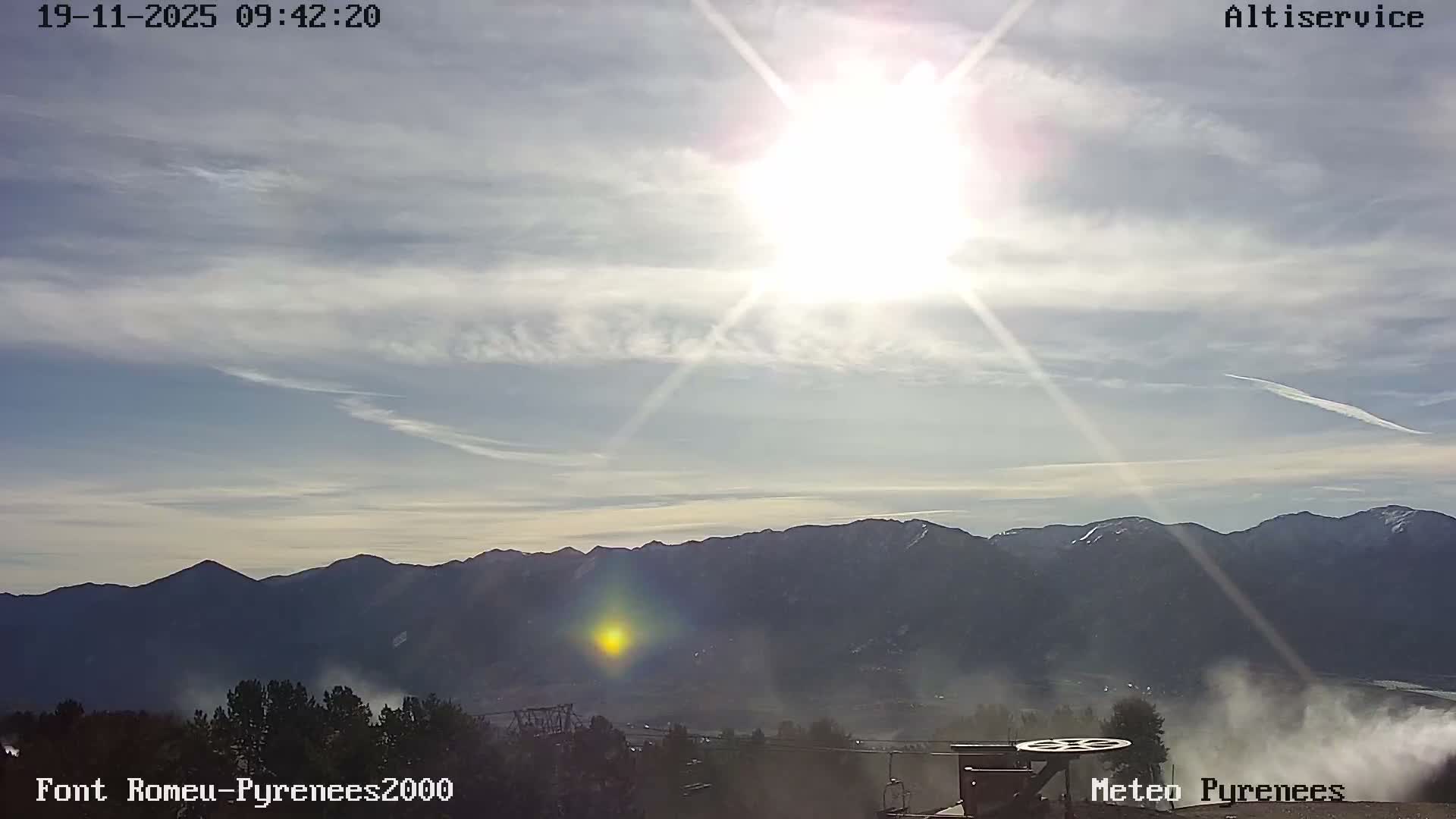 Under a bright, sunny sky with scattered wispy clouds and visible contrails, a panoramic view reveals a range of dark mountains with patches of snow, overlooking a misty valley with evergreen trees and a ski lift in the foreground.
