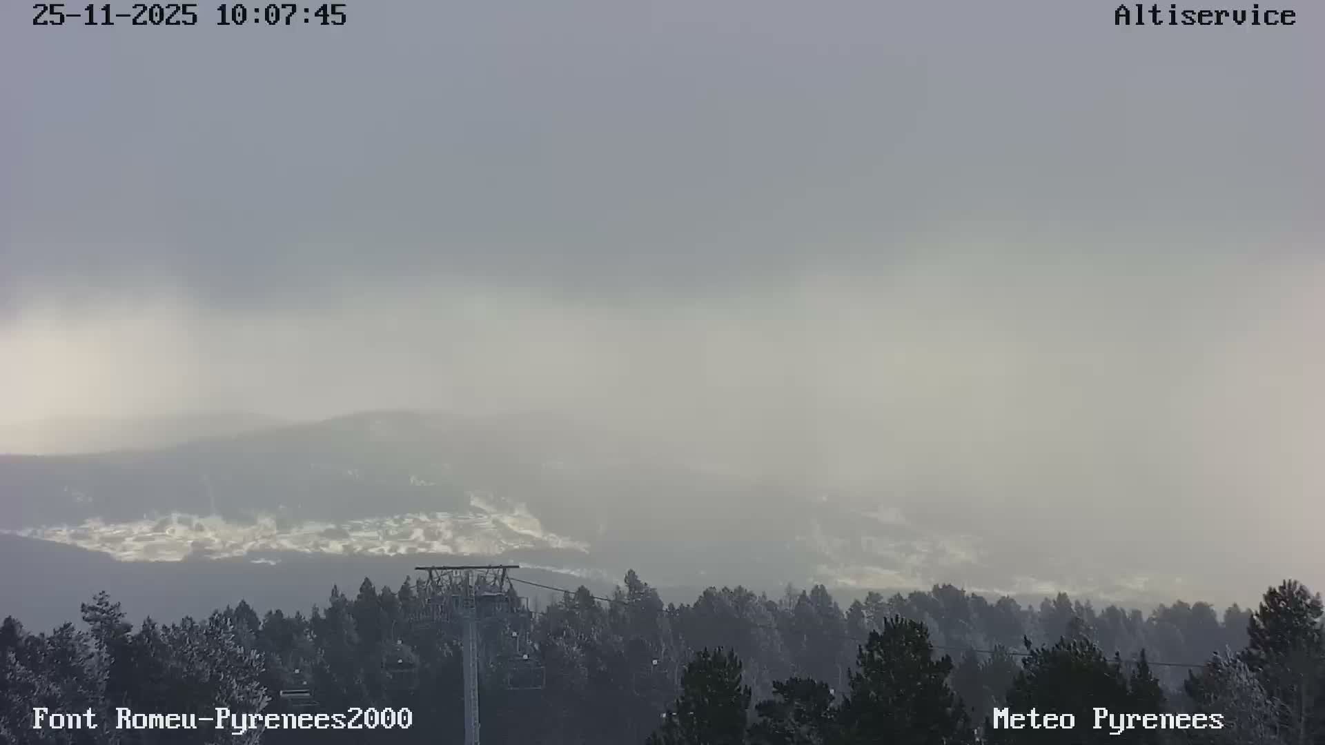 A winter mountain scene shows snow-dusted pine trees and a ski lift in the foreground, with distant peaks largely obscured by heavy fog and an overcast sky.