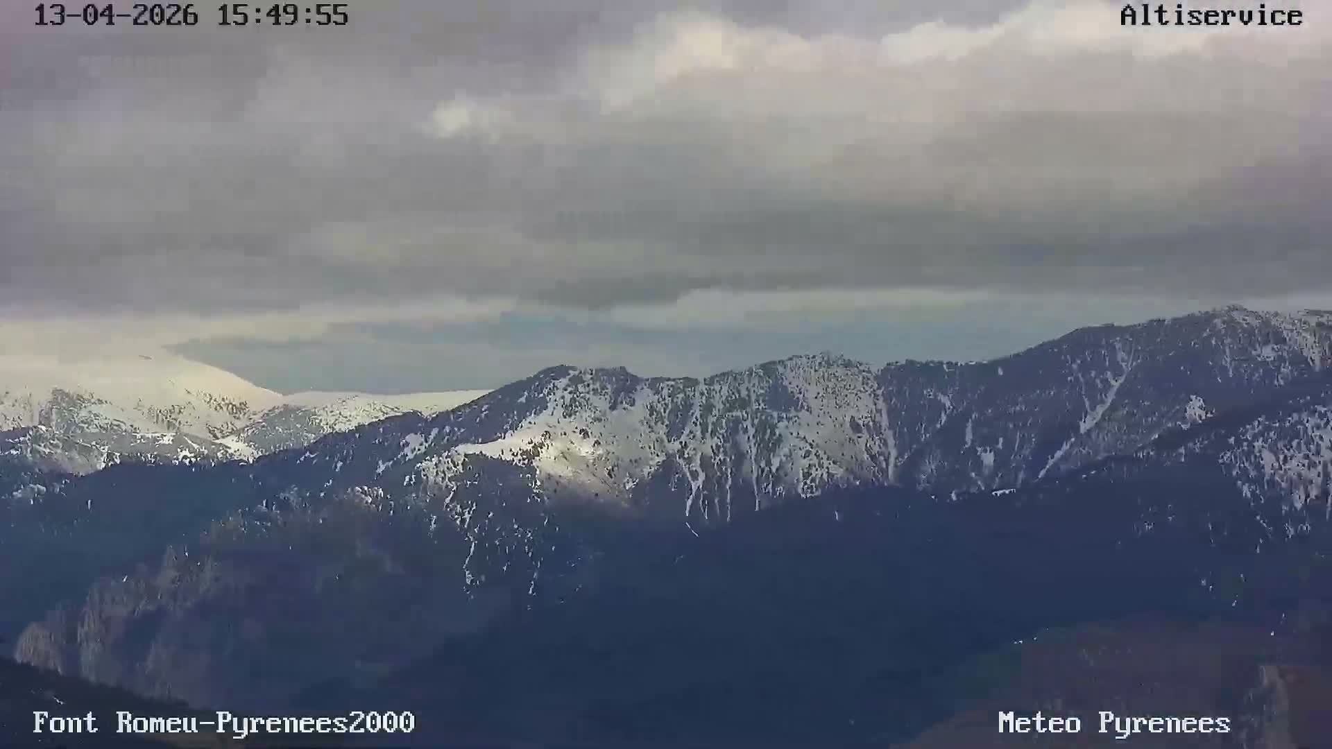 A winter mountain scene shows snow-dusted pine trees and a ski lift in the foreground, with distant peaks largely obscured by heavy fog and an overcast sky.