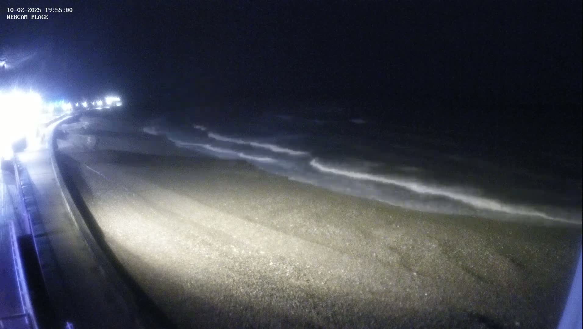 A dark, nighttime beach scene shows small waves lapping a pebbled shore next to a roadway with lights visible in the distance.