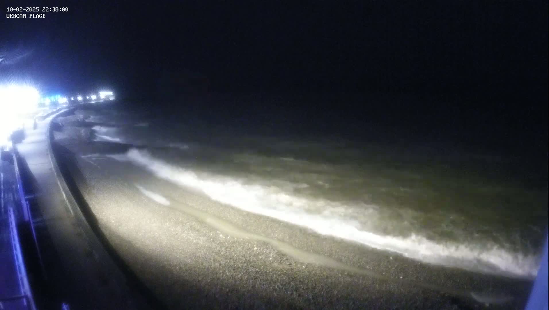 At night, waves crash on a pebbled shore next to a paved walkway.