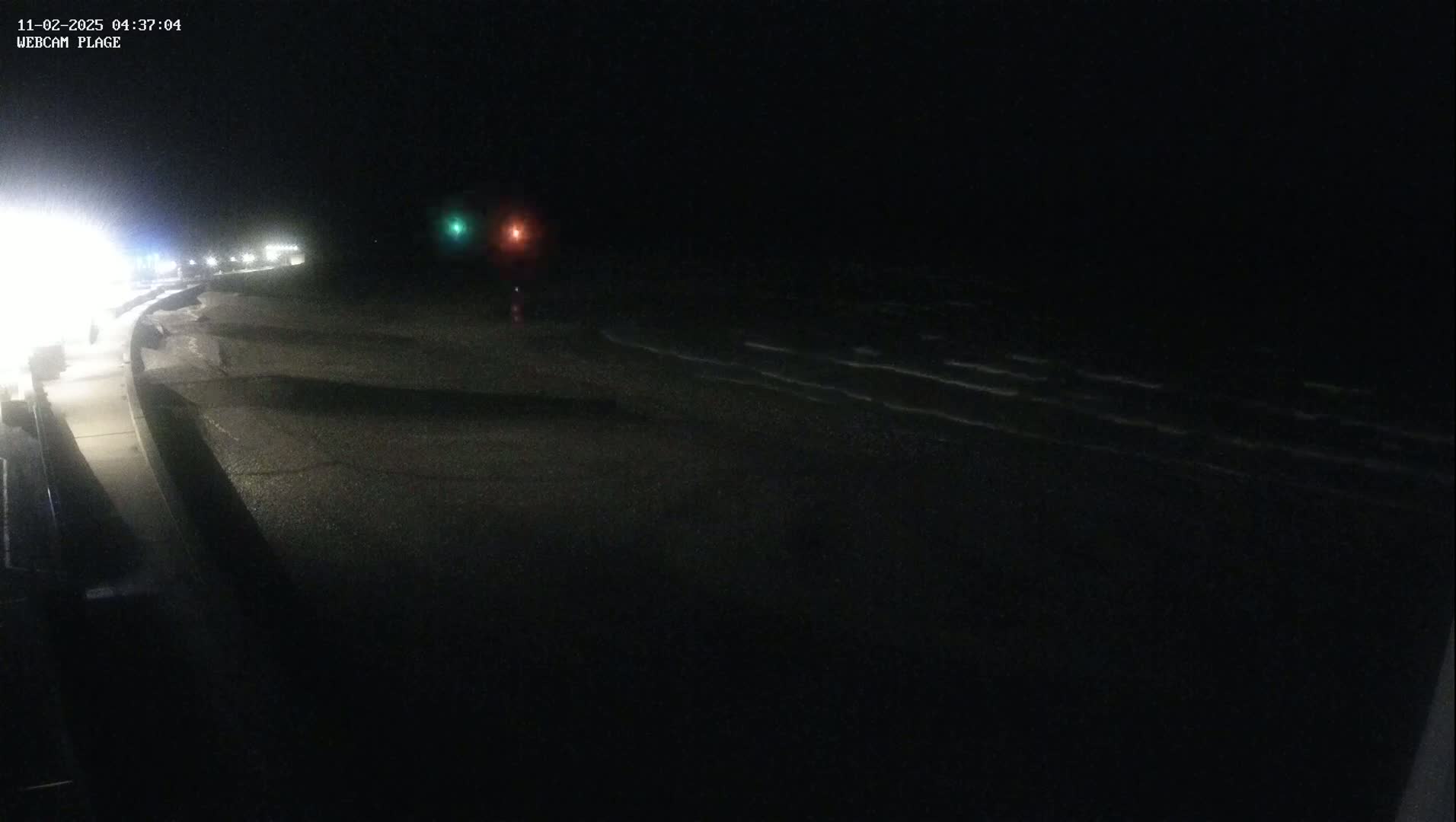 A dark, nighttime beach scene shows gently lapping waves on a shore with a paved walkway and distant lights visible.