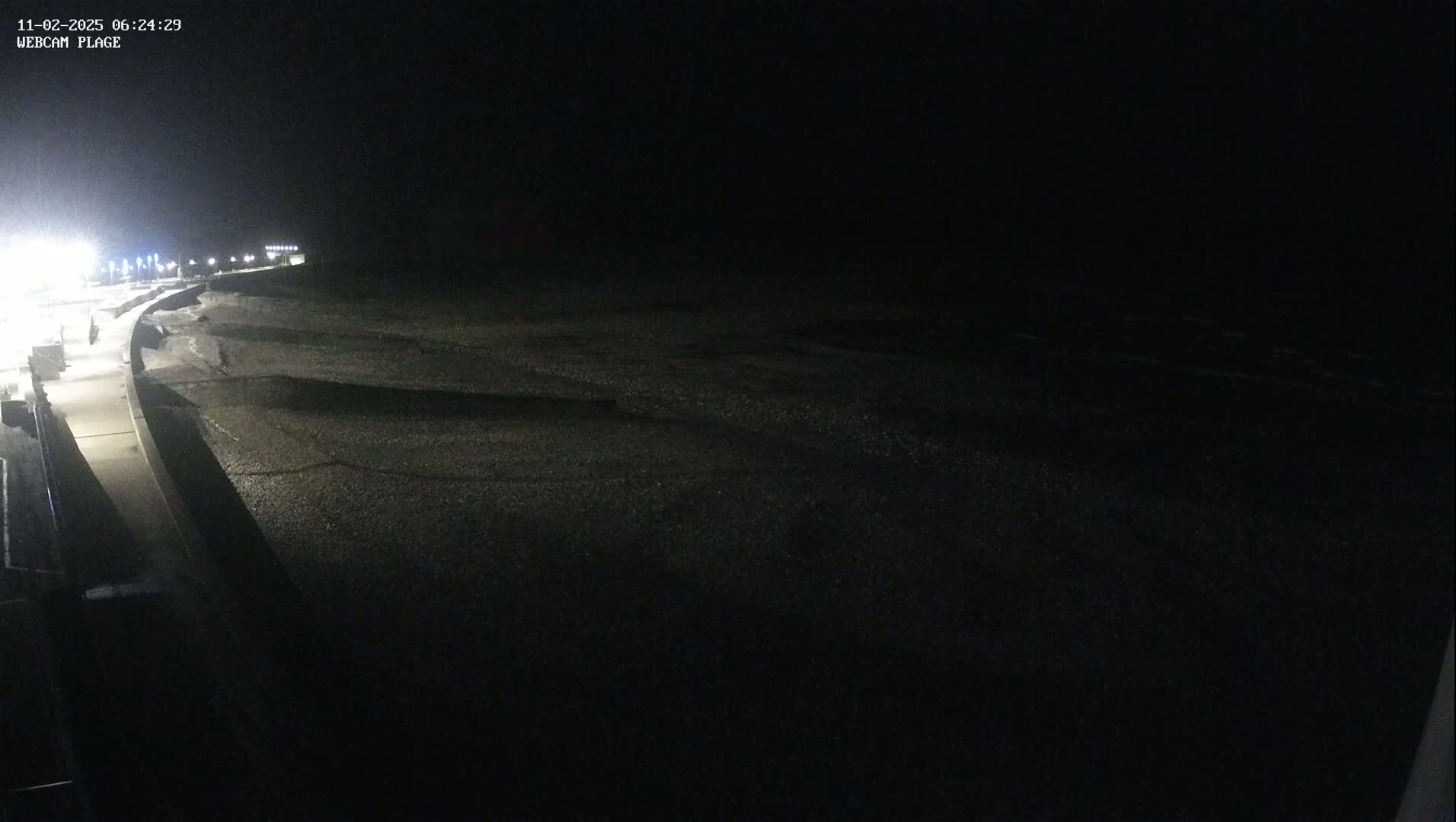 A dark, nighttime view shows a sparsely lit walkway next to a dark beach and ocean under a clear, starless sky.