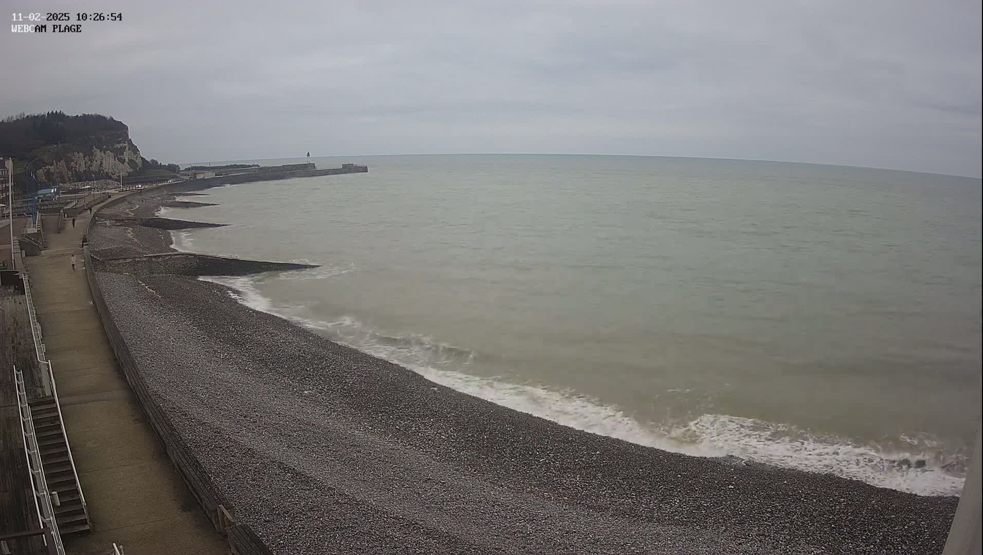 A pebble beach with gentle waves rolls onto shore under an overcast sky, next to a paved walkway and a cliff in the distance.