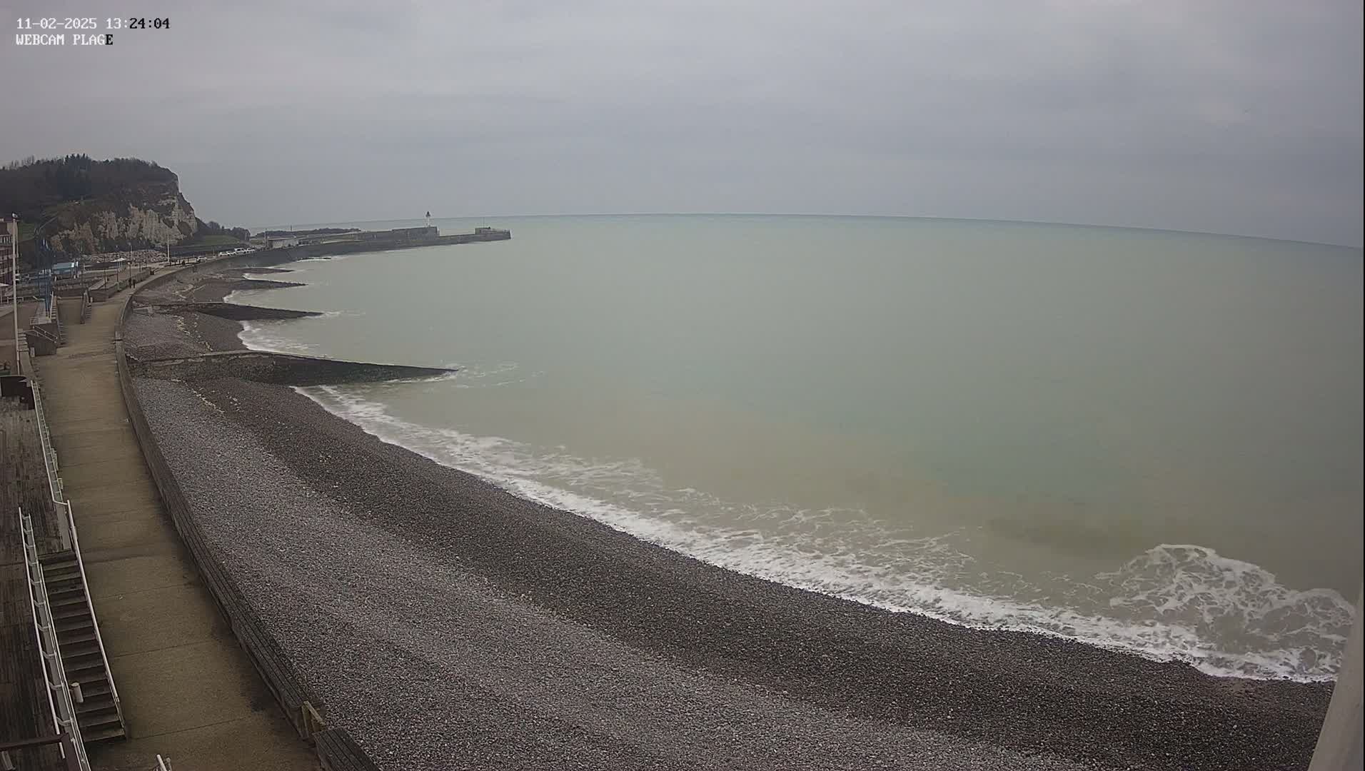 A pebbled beach and walkway are shown next to a calm, overcast sea, with a distant cliff in the background.
