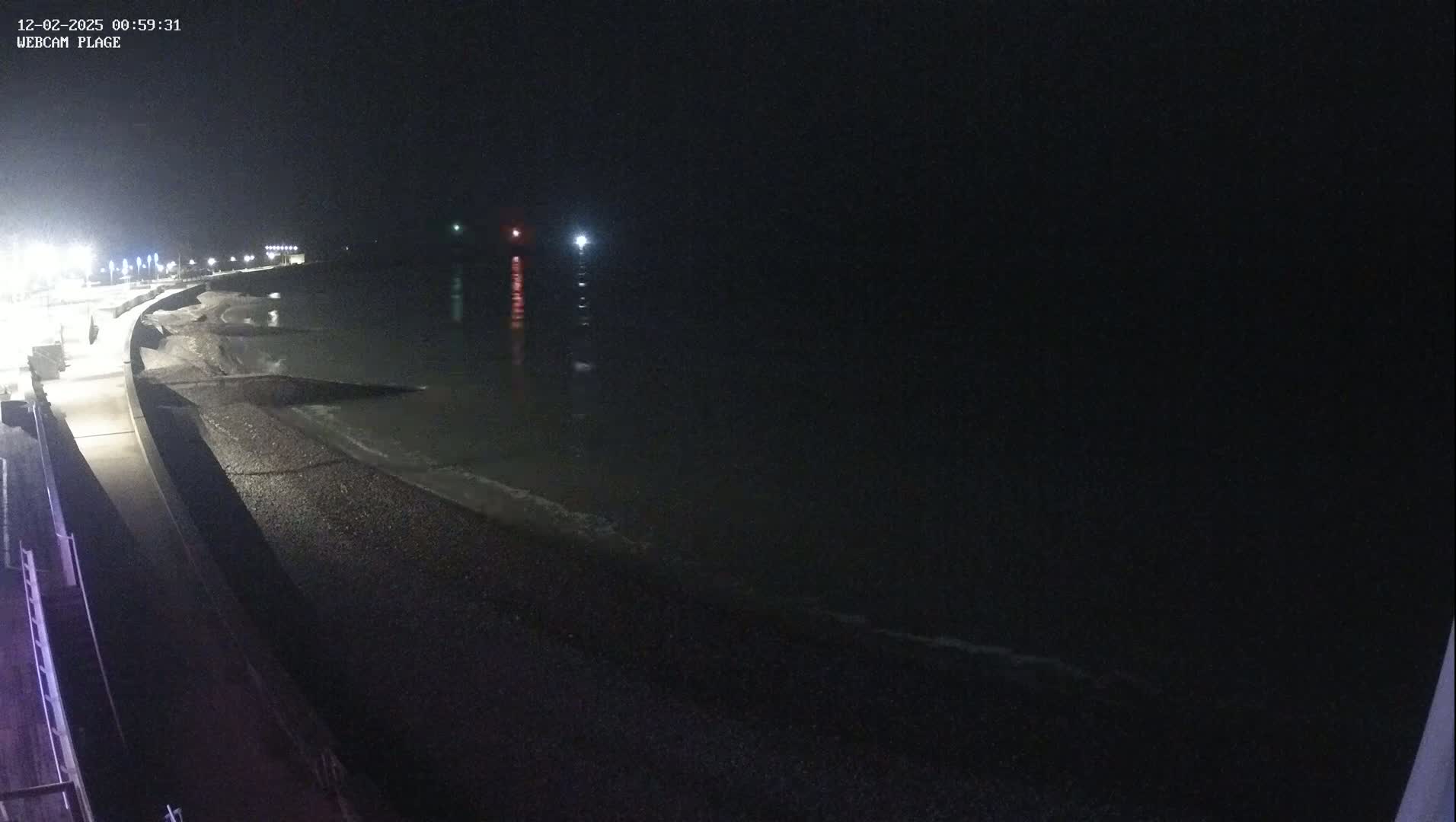 A dark, nighttime view shows a pebbled beach and calm ocean waves lapping the shore next to a paved walkway lined with lights.