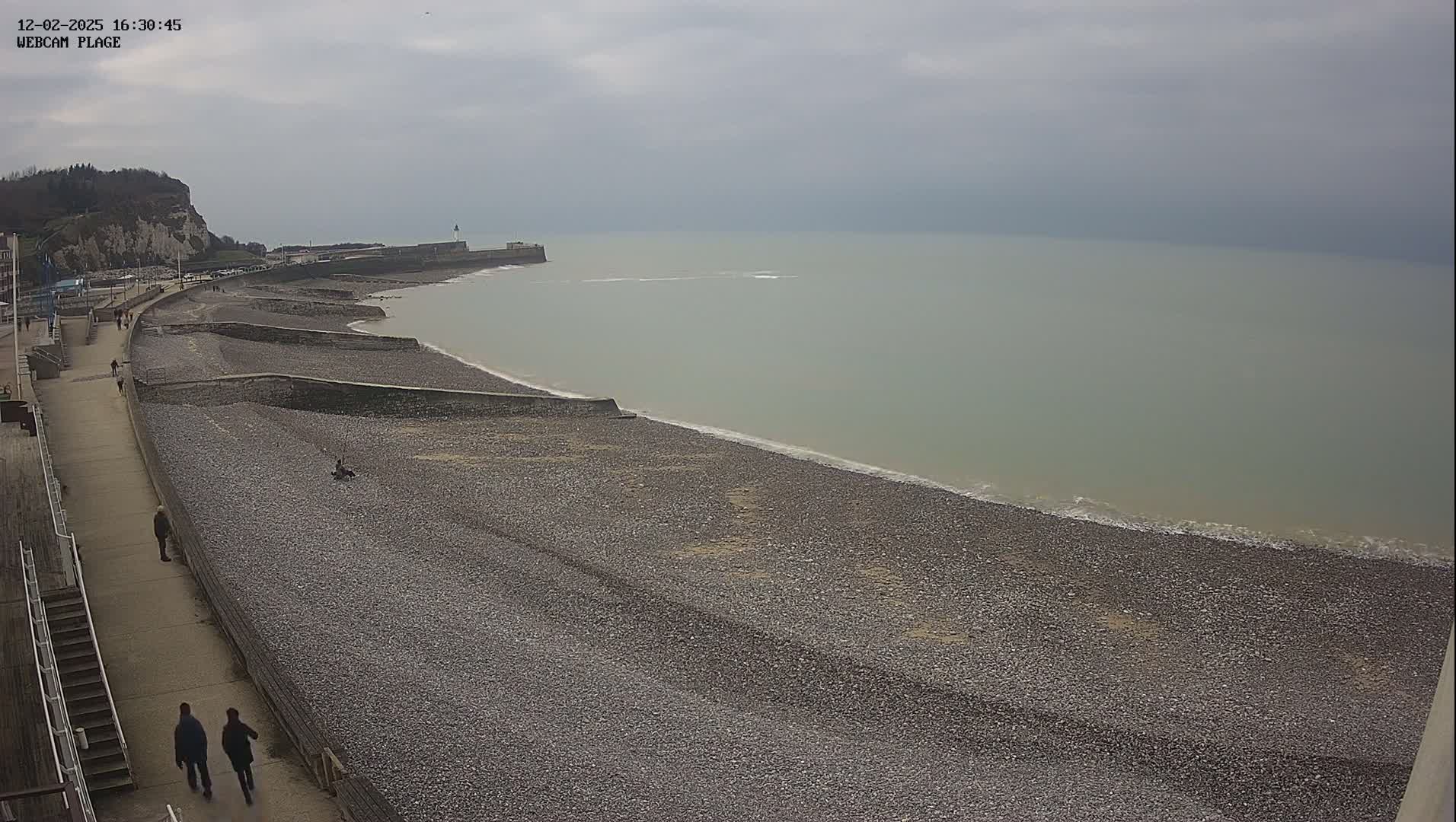 A long, pebbled beach stretches along a calm, gray sea under an overcast sky, with a few people walking along a promenade beside it.