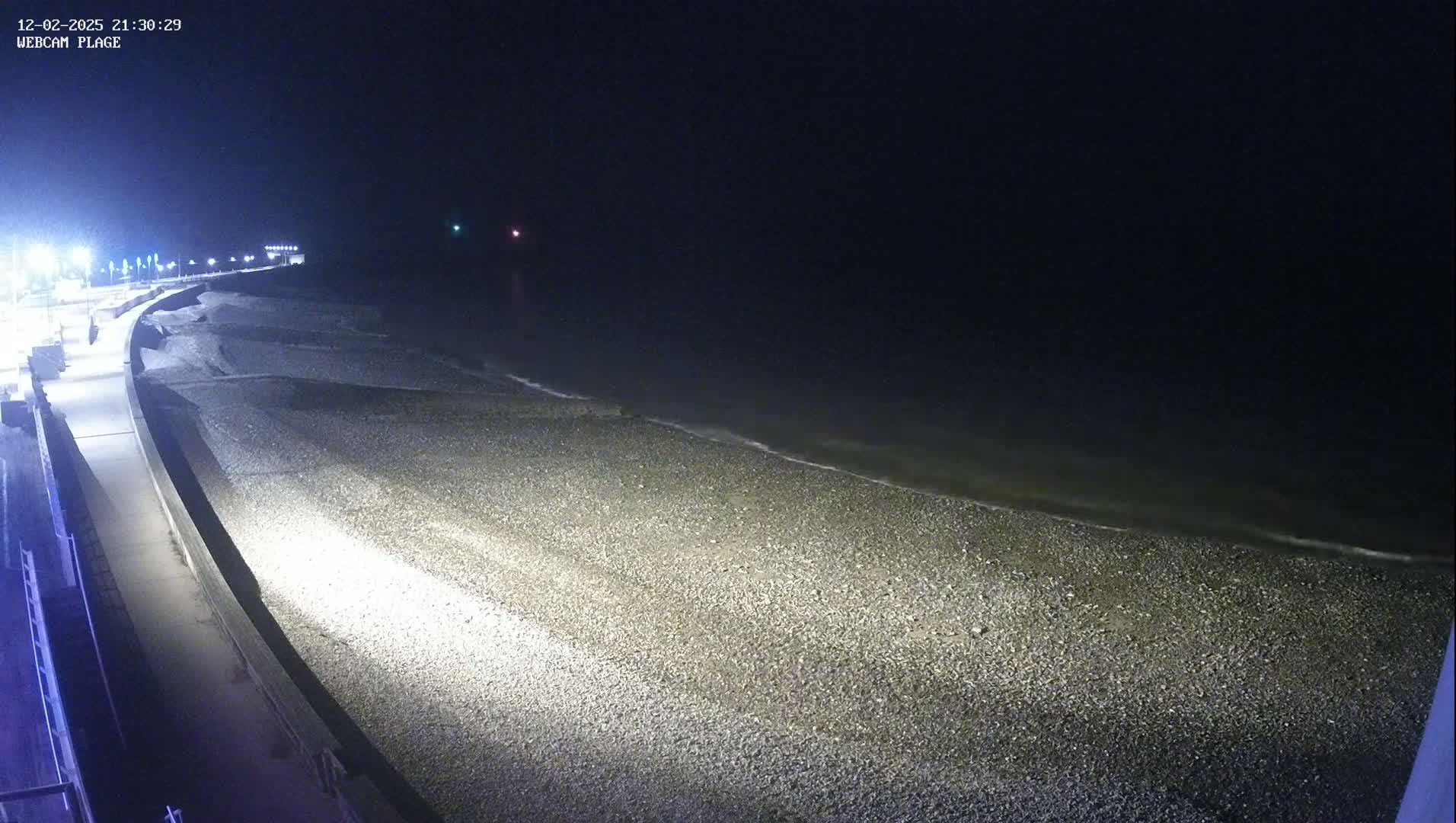 A nighttime view of a pebble beach illuminated by artificial light, with a calm, dark ocean visible in the background.