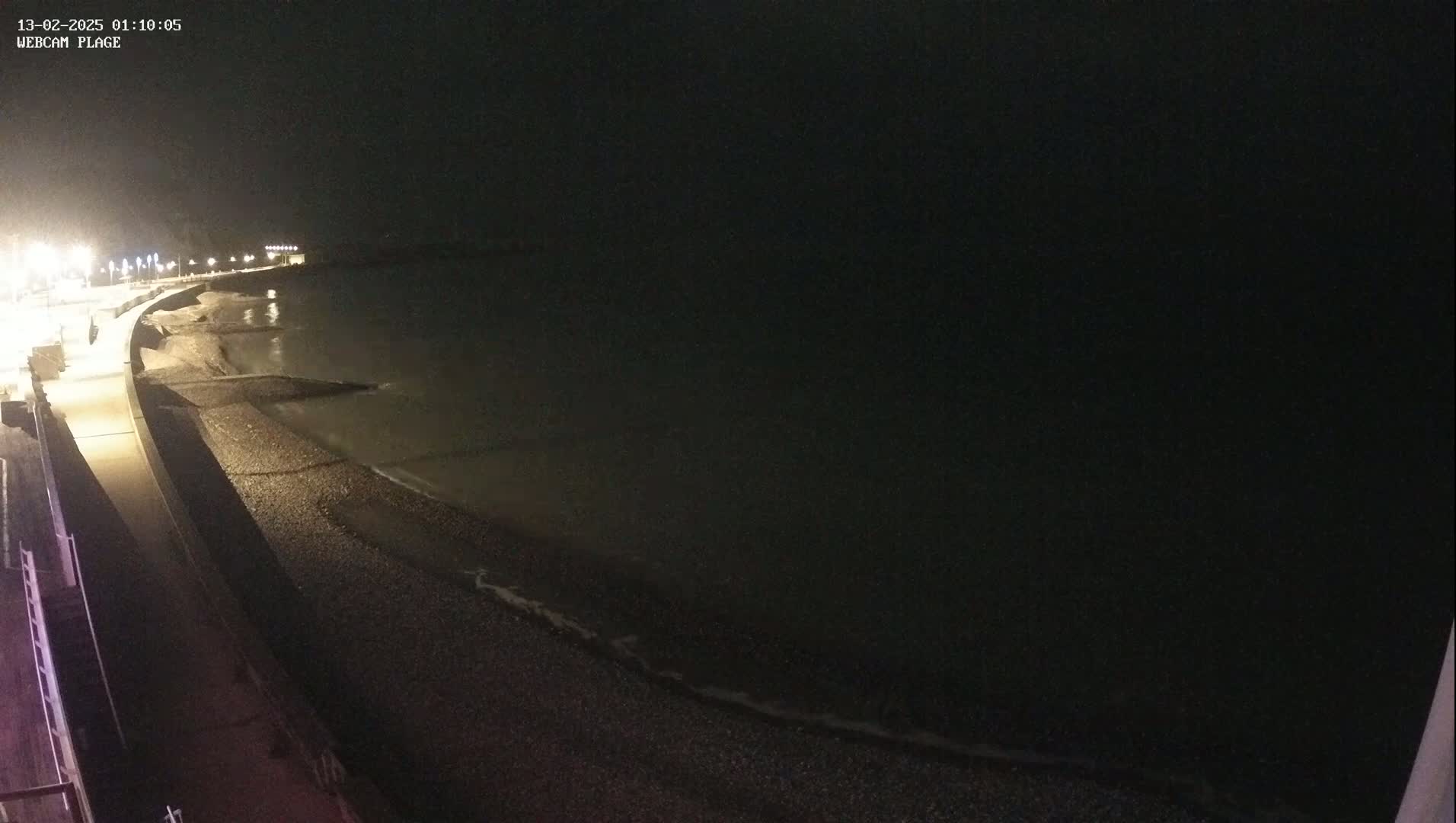 A dark, nighttime view of a pebble beach and a paved walkway beside a calm sea, illuminated by streetlights.
