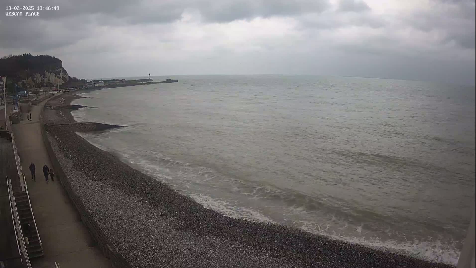A gray, overcast day at a pebble beach shows a calm sea with small waves lapping the shore, a walkway with a few people walking, and a cliff in the background.