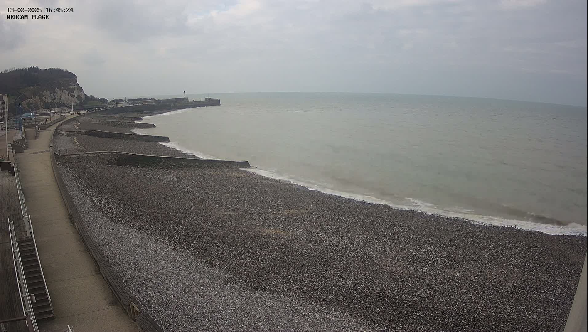 A pebble beach stretches along a calm sea under an overcast sky, with a paved walkway and a cliff visible in the background.