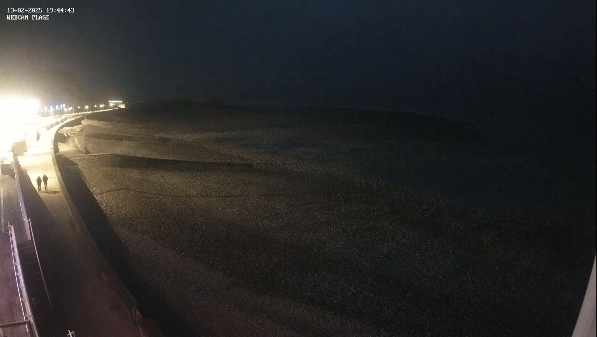 Two people walk along a lit pathway next to a dark, pebble-covered beach at night.