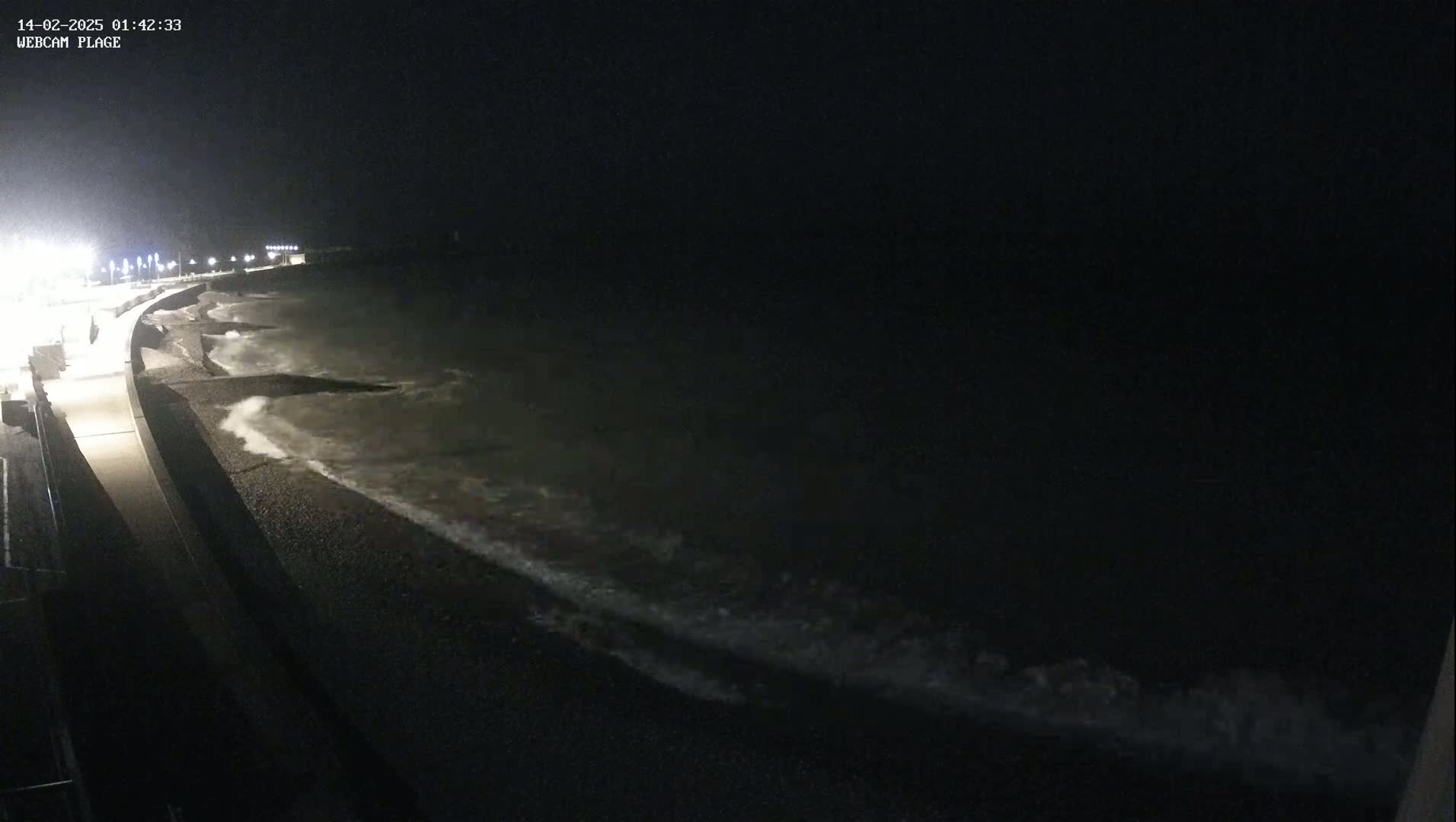 At night, waves crash on a dark, pebbled shoreline next to a lit walkway.