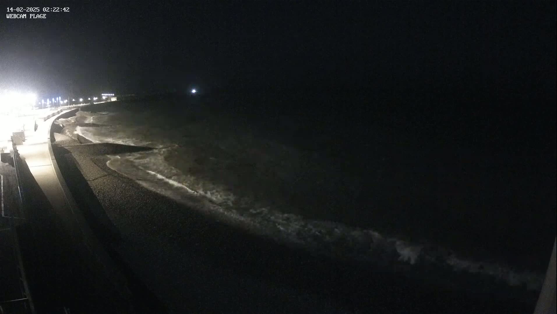 Waves crash on a dark, pebbled shoreline at night, next to a lit walkway.