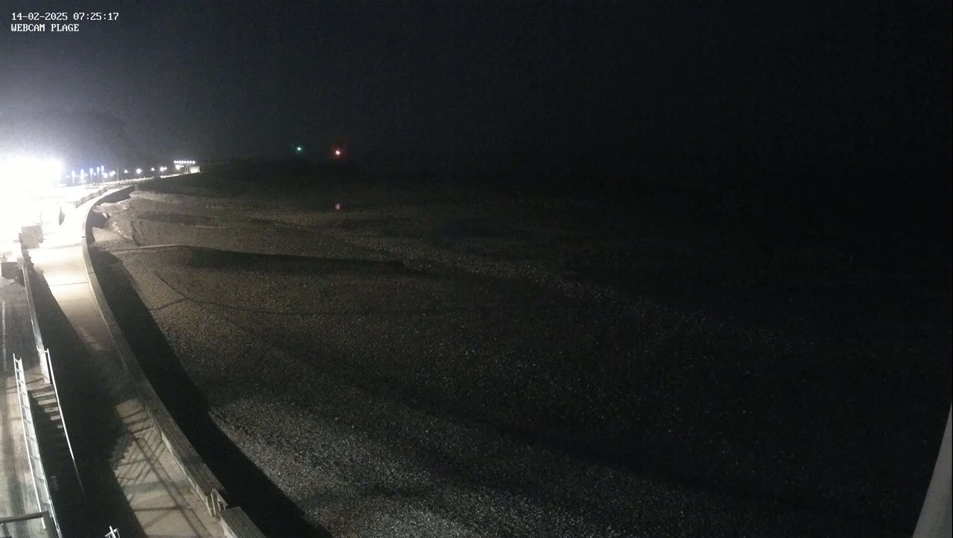 A dark, nighttime view of a pebble beach and a paved walkway, under a mostly cloudy sky.
