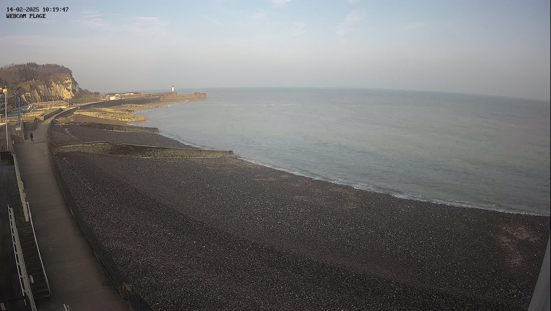 A mostly clear day reveals a pebble beach stretching to a calm sea, where a walkway leads past a cliff to a distant lighthouse.