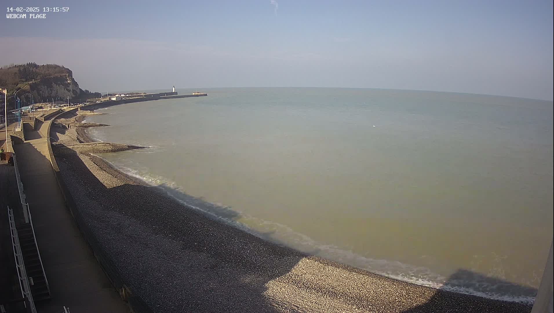 A pebble beach curves along a calm, light-brown sea under a clear blue sky, with a distant cliff and a breakwater visible.