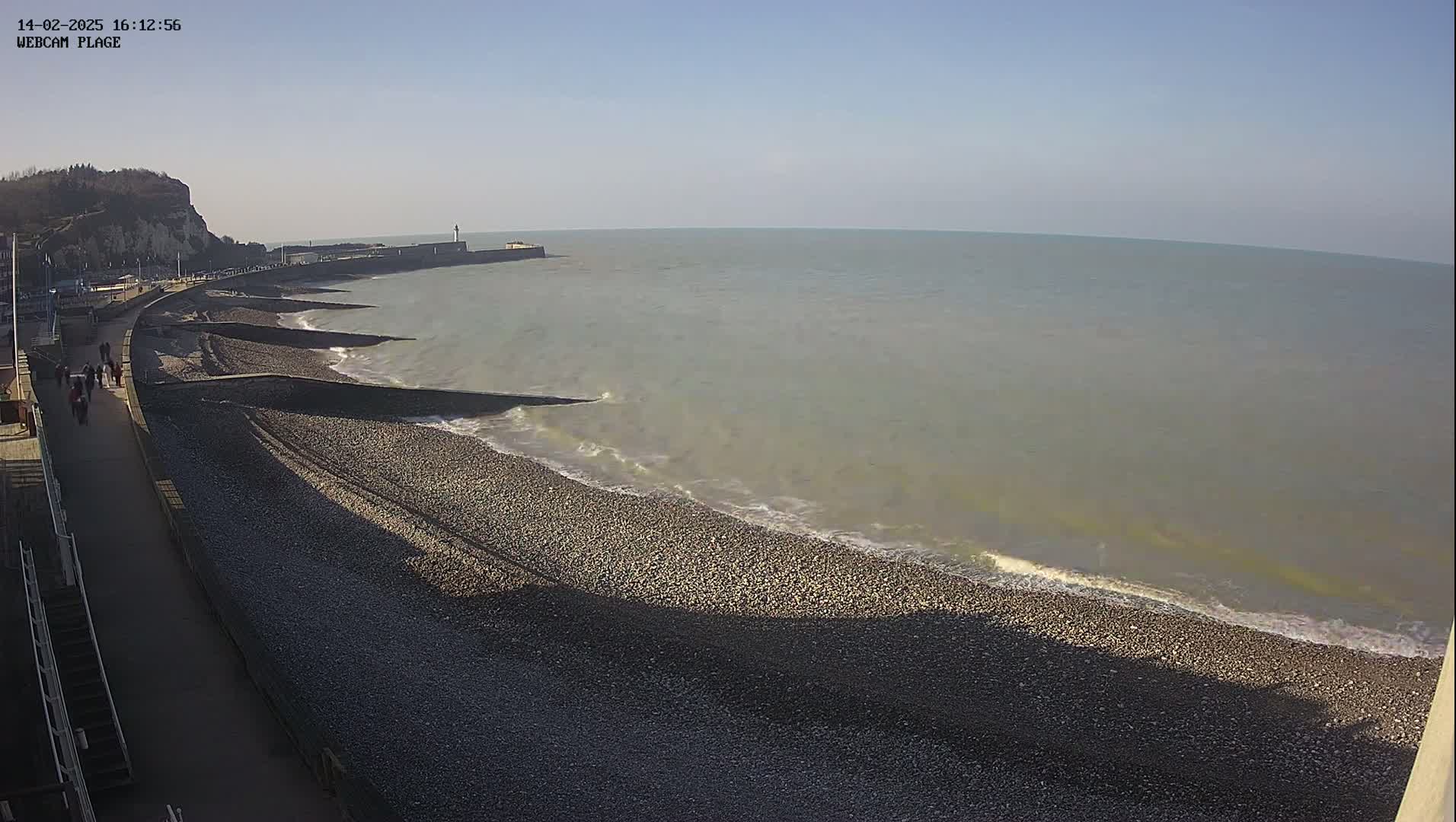 A pebble beach and a walkway are seen alongside calm, light-grey water under a clear blue sky.