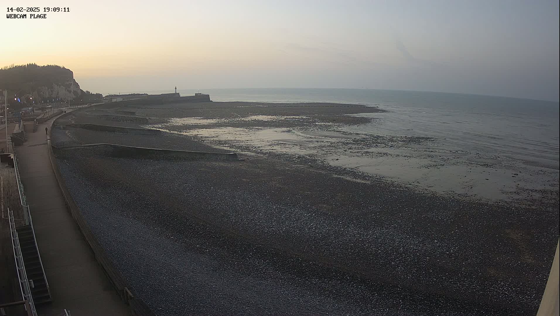 A wide shot of a pebble beach at low tide under a dusky sky shows a paved walkway leading away from the shore toward a cliffside.