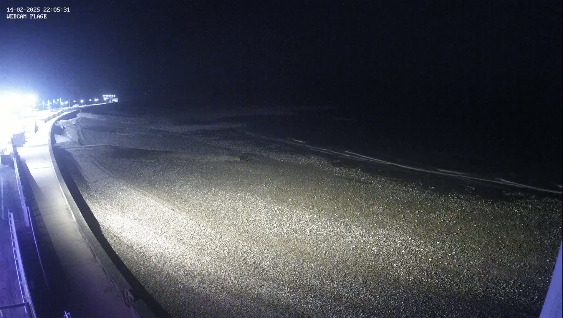 A dark, nighttime view of a pebble beach and a paved walkway, illuminated by artificial lights, with a calm, dark ocean in the background.