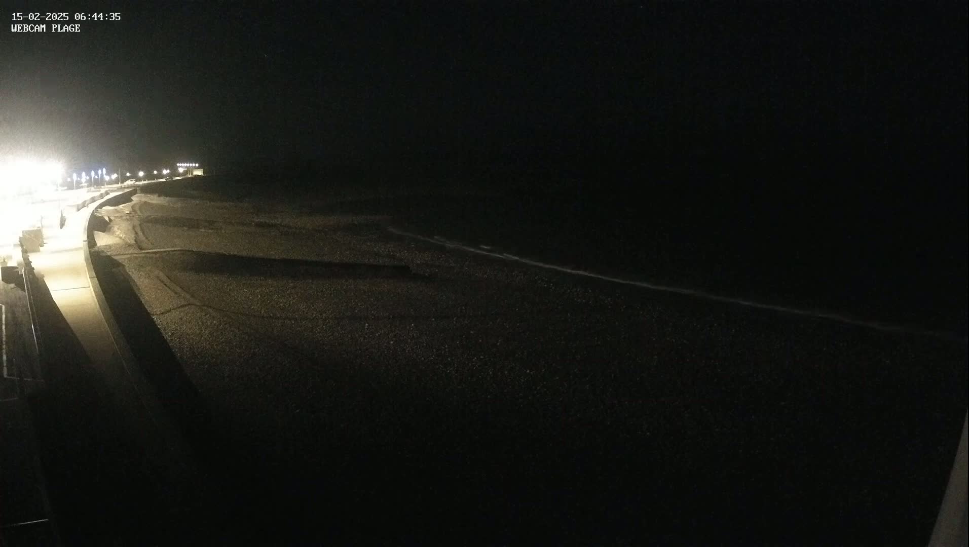 A dark, nighttime view of a pebble beach and a lit promenade, with a calm, dark ocean in the background.