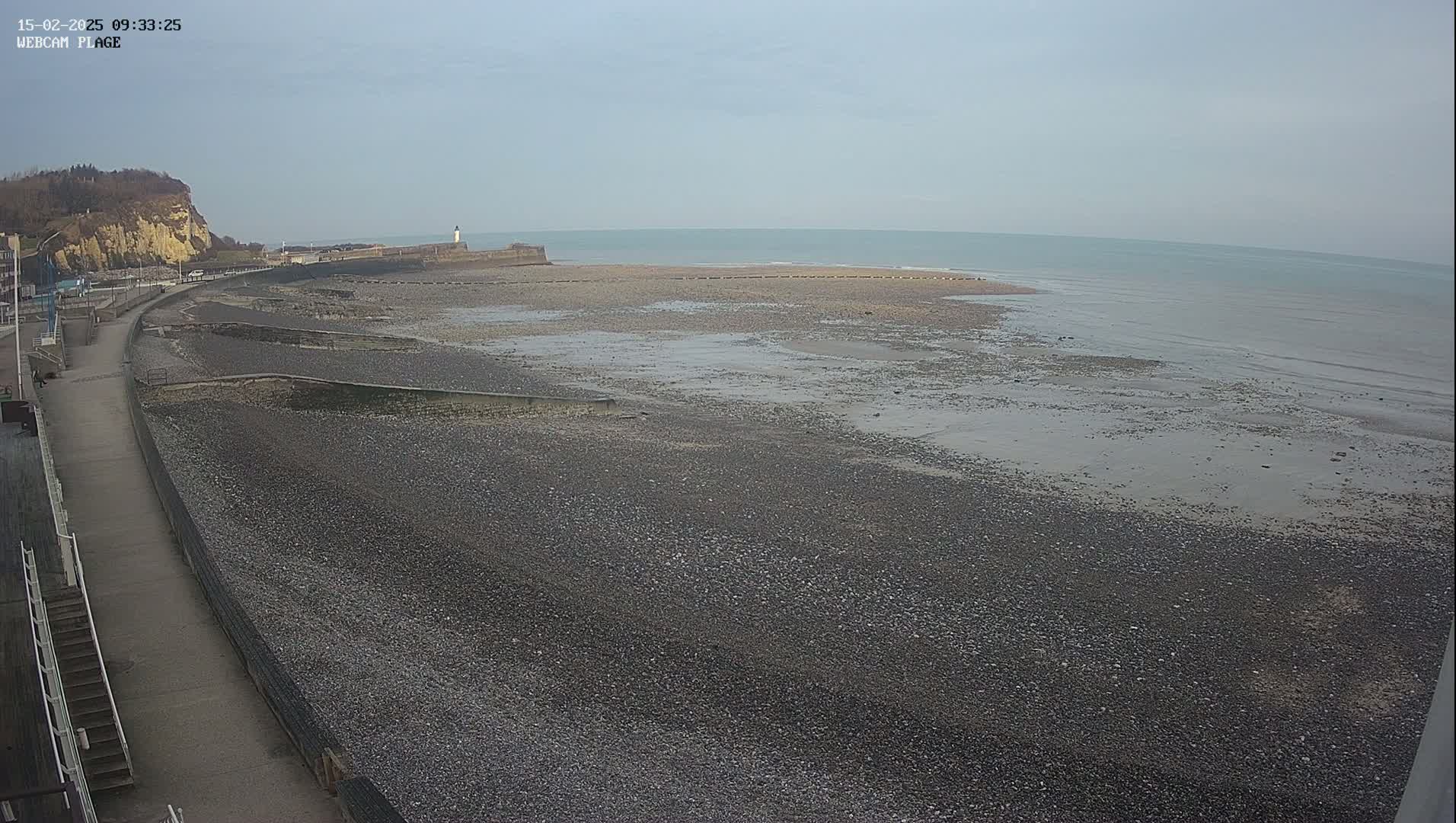 A mostly gray, pebble beach stretches out to a calm, gray-blue sea under a cloudy sky, with a paved walkway and a cliff visible in the background.