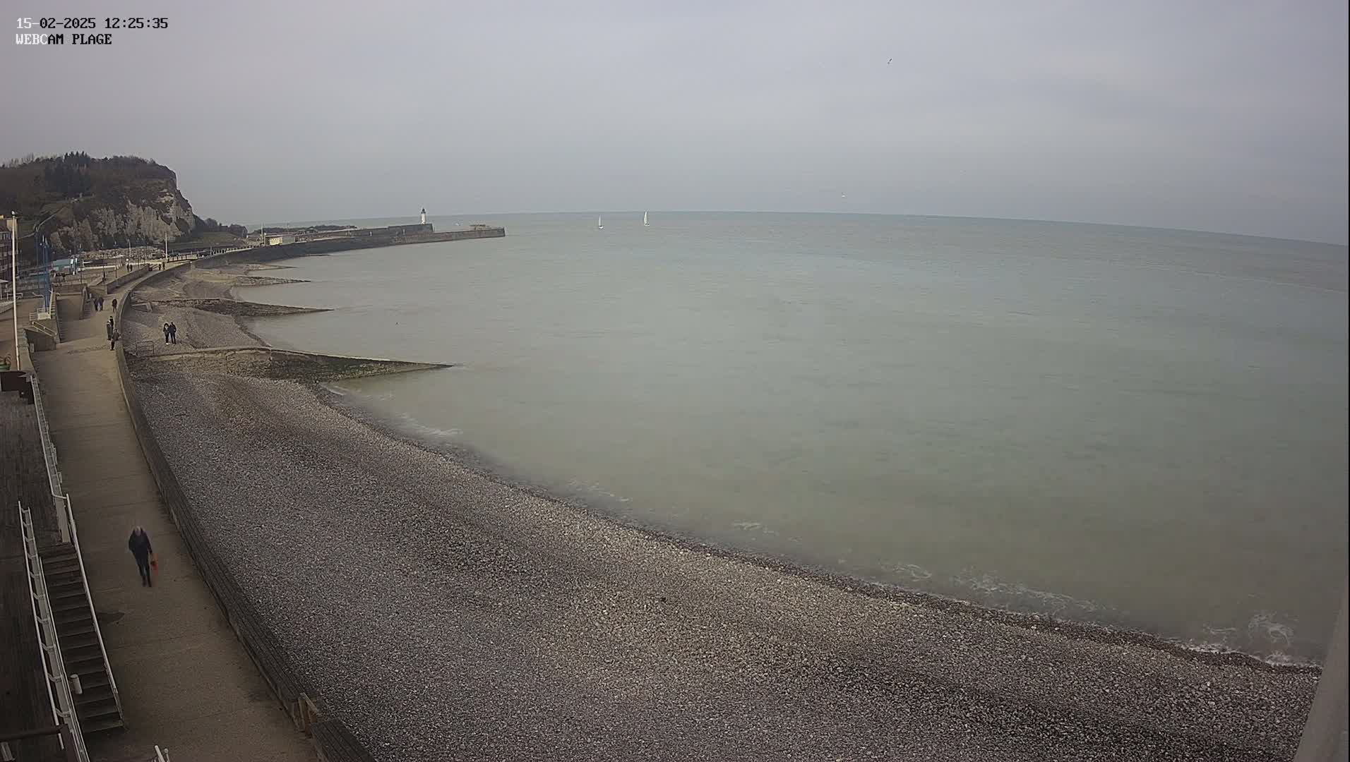 A pebble beach stretches along a calm, grey sea under an overcast sky, with a few people walking on the promenade and a distant lighthouse visible.