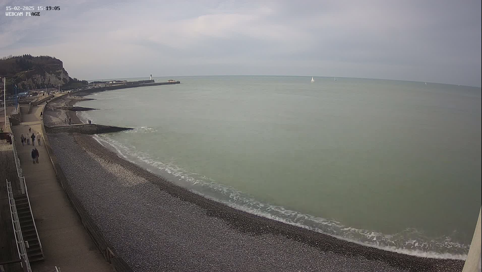 A pebble beach with a walkway and several people walking along it, meets a calm, light-green sea under an overcast sky.