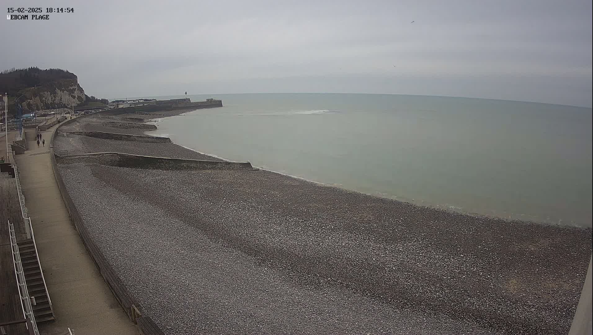 A pebble beach stretches along a calm, gray sea under an overcast sky, with a walkway and buildings visible in the background.
