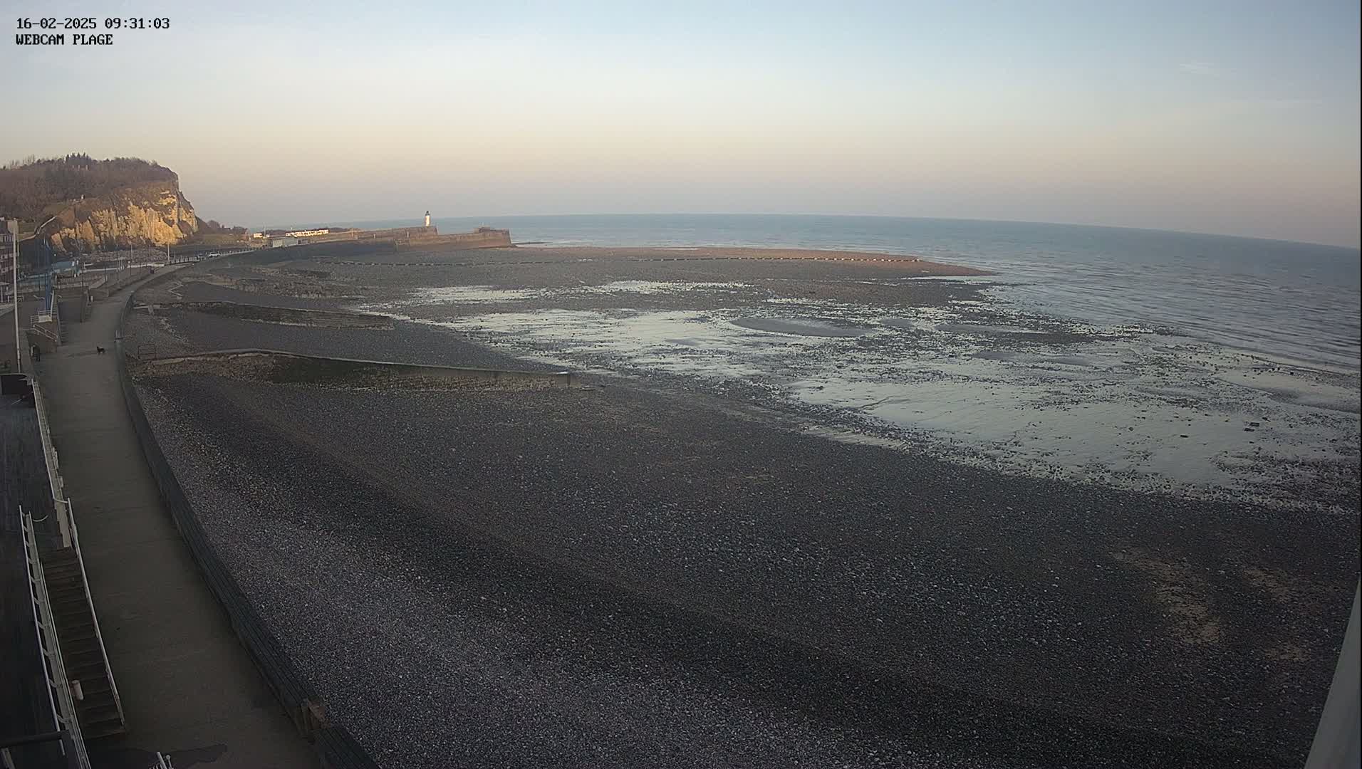 A mostly clear, twilight sky overlooks a dark, pebbled beach and calm ocean at low tide, with a paved walkway and a cliff visible in the background.