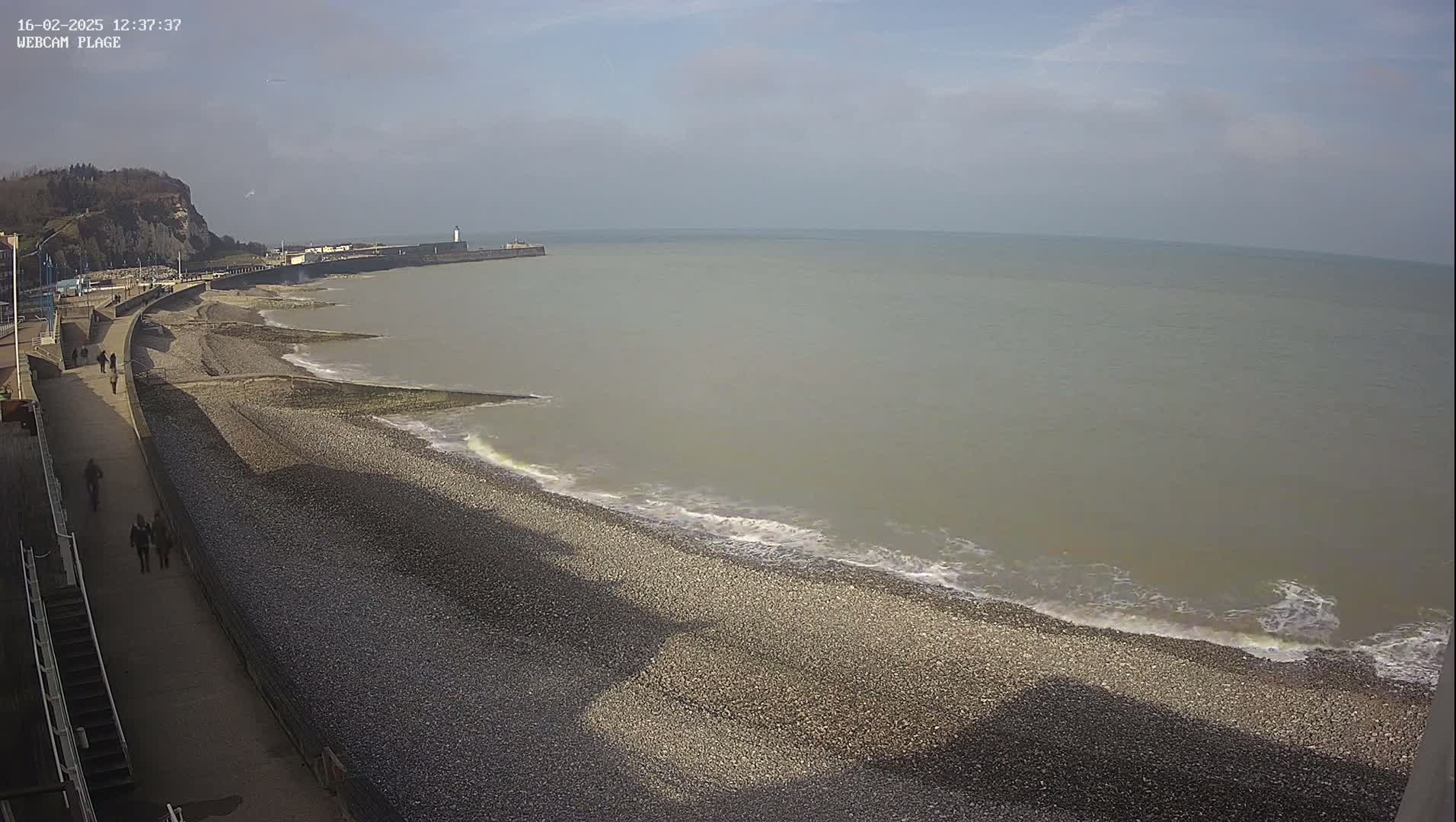 A pebble beach stretches along a sea wall under a mostly clear sky, with gentle waves lapping at the shore and a few people walking along the promenade.