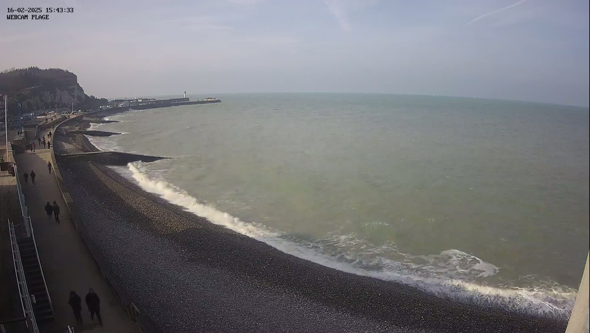 A rocky beach with a walkway and people walking along it, with moderate waves breaking on the shore under a partly sunny sky.