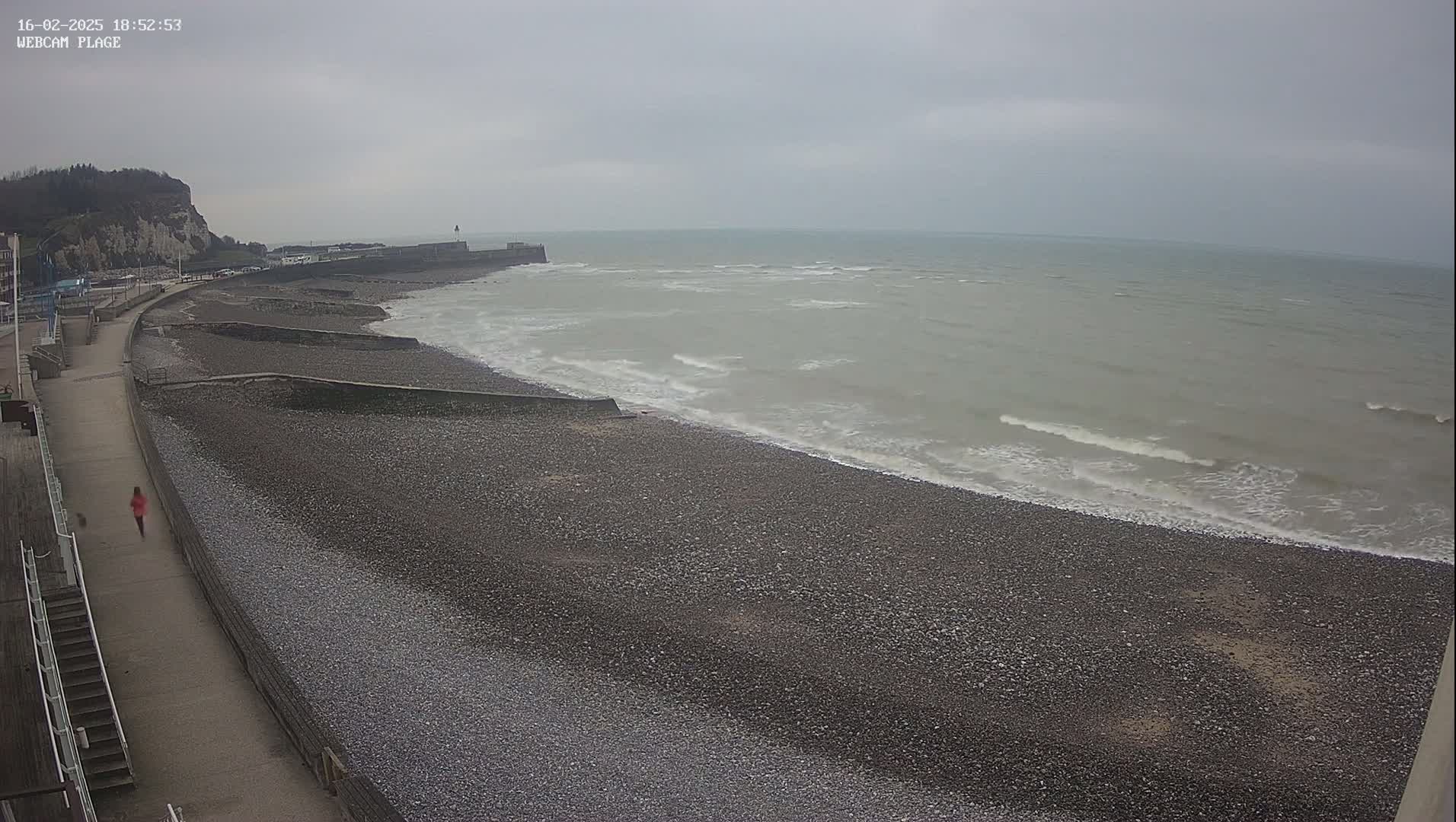 A person walks along a paved path next to a pebble beach where small waves lap the shore under an overcast sky.