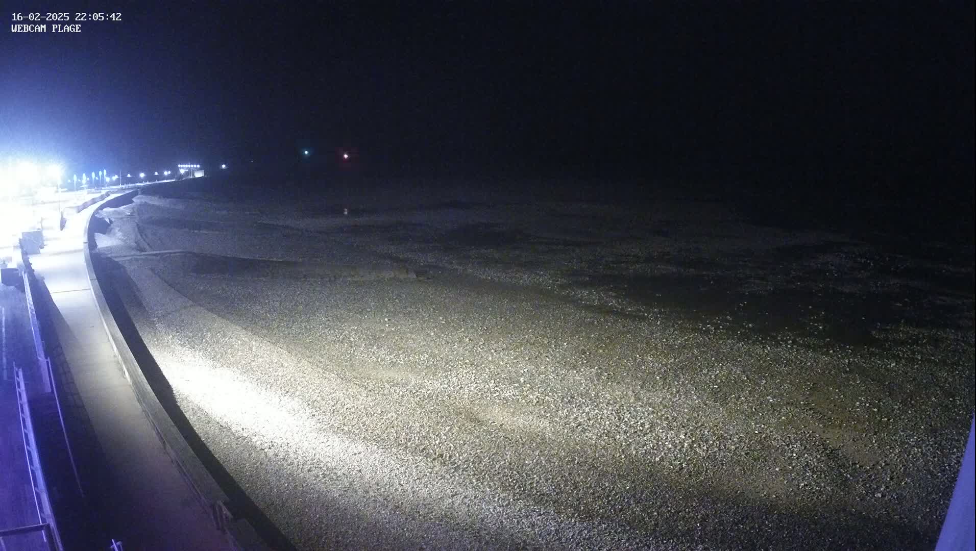 A dark, nighttime view shows a pebbled beach illuminated by artificial light, with a paved walkway curving along the shoreline.