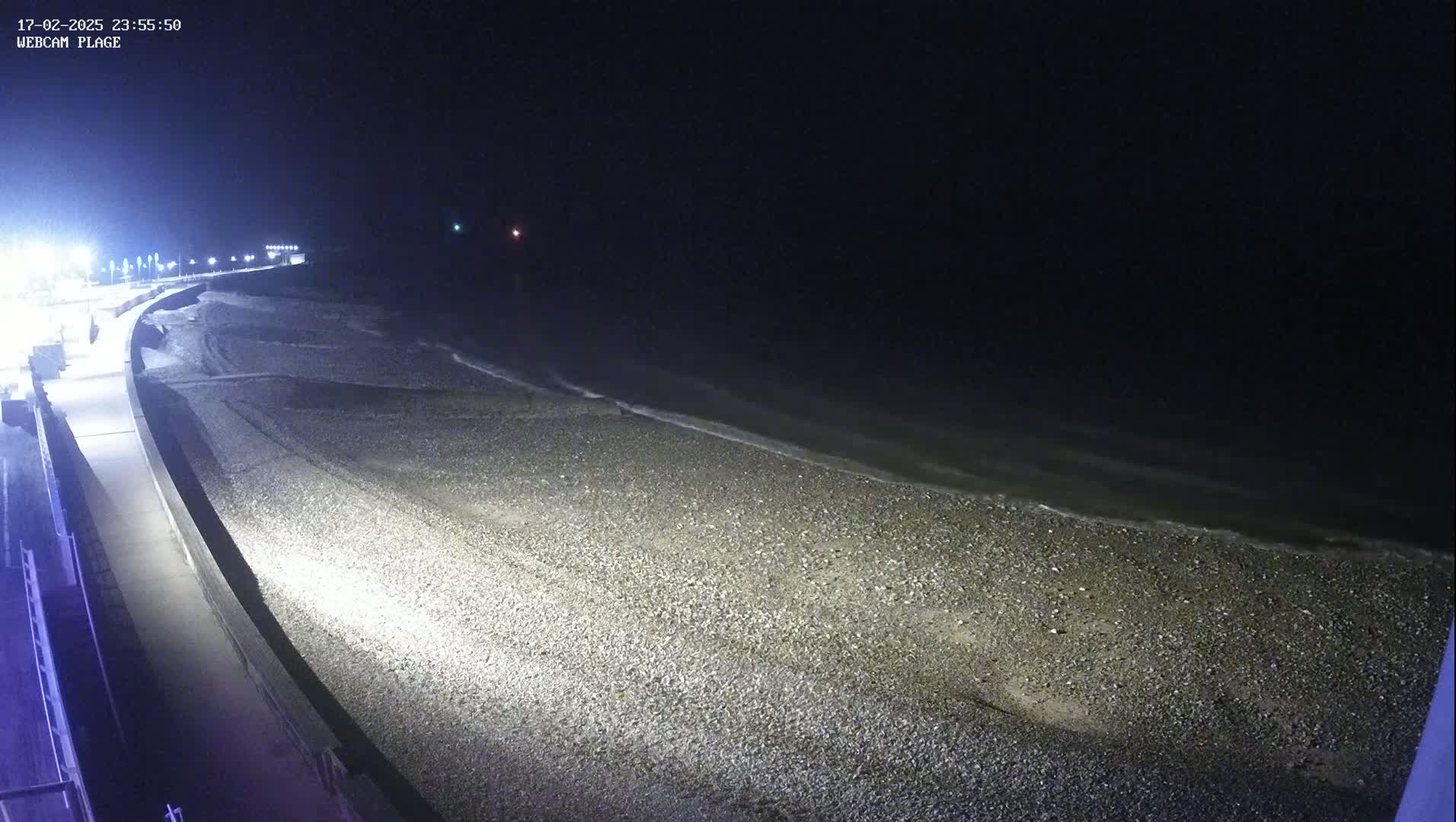 A dark, pebble beach is gently lapped by waves at night, illuminated by streetlights along a nearby walkway.