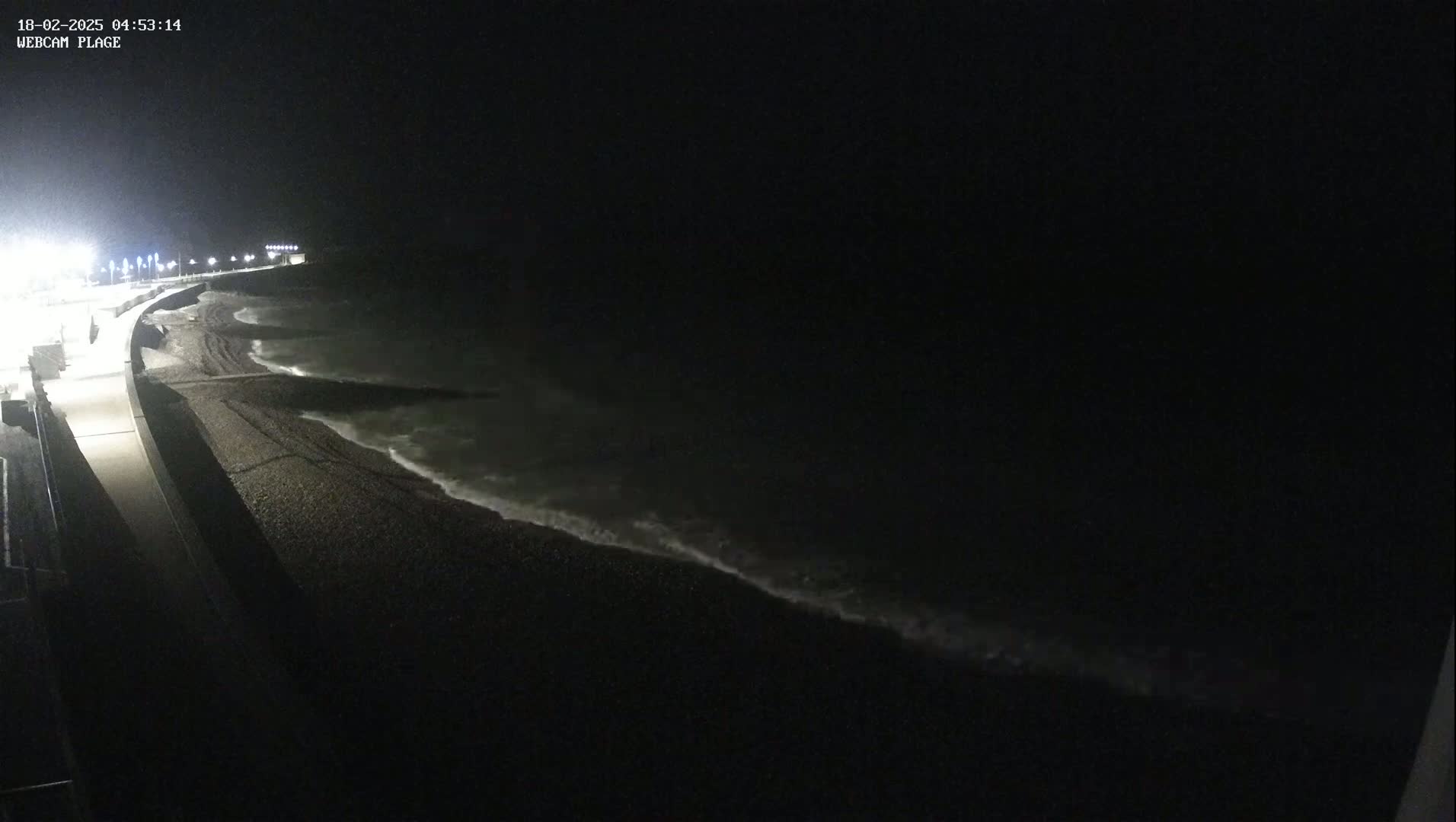 A dark, nighttime view shows a pebble beach and ocean waves lapping the shore under a clear, black sky, with a lit walkway visible in the foreground.