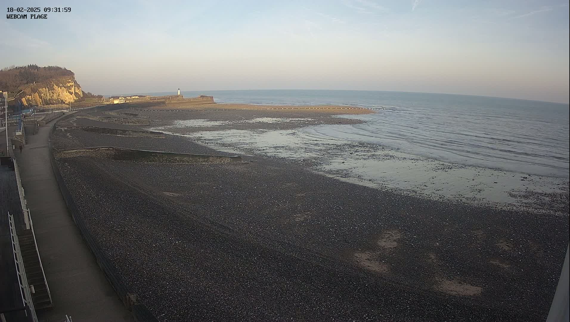 A mostly clear, daytime view of a pebble beach and calm ocean, with a paved walkway and a cliff in the background.