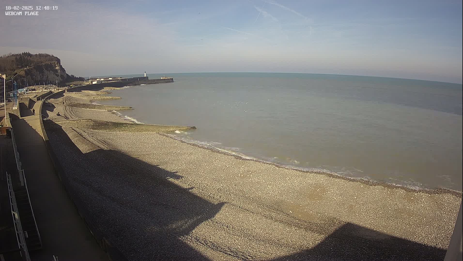 A pebble beach stretches along a calm sea under a mostly sunny sky, with a cliff and a walkway visible in the foreground.