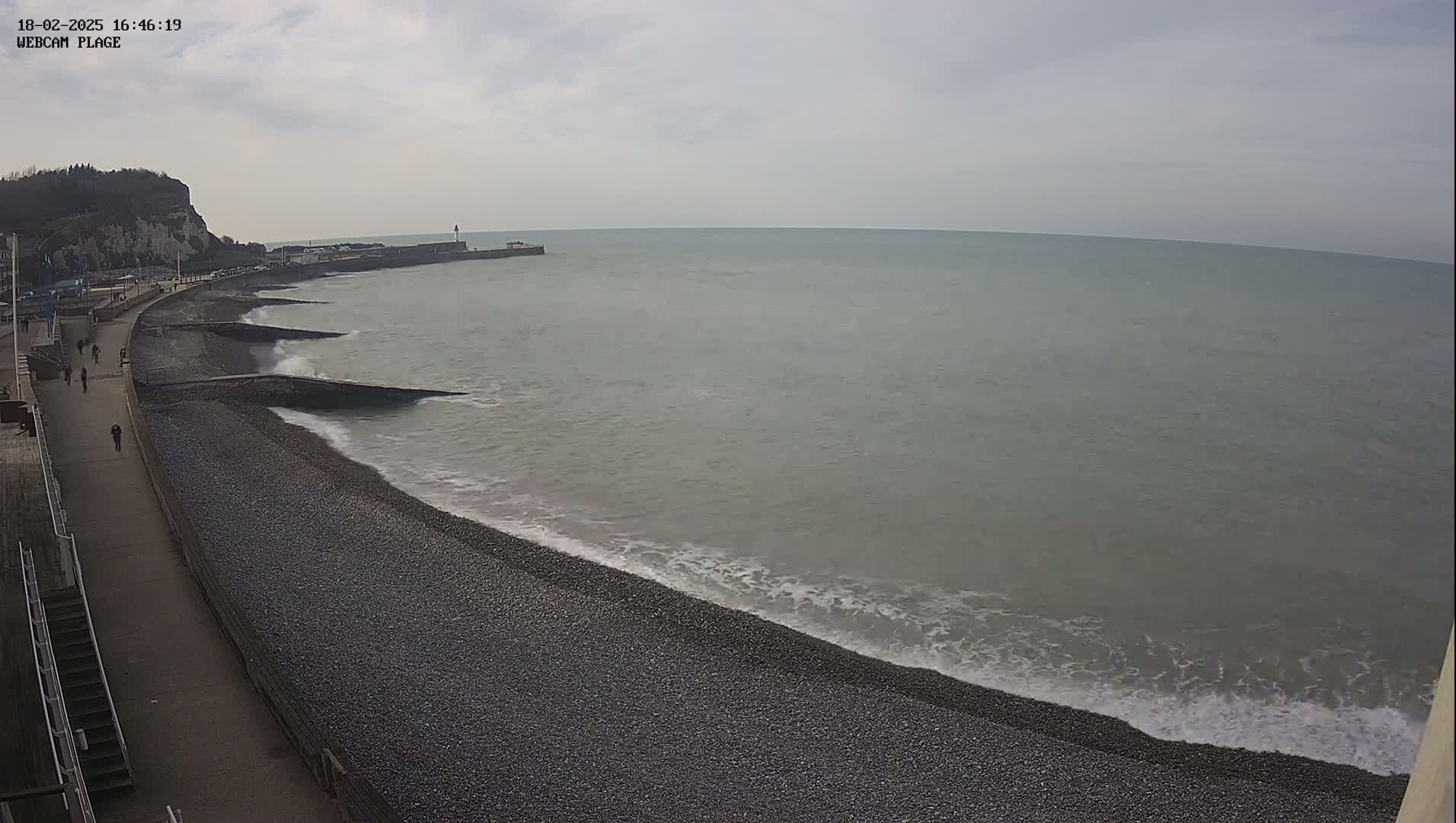 A pebble beach with a walkway and several people walking on it stretches along a calm, grey sea under an overcast sky, with a cliff visible in the distance.