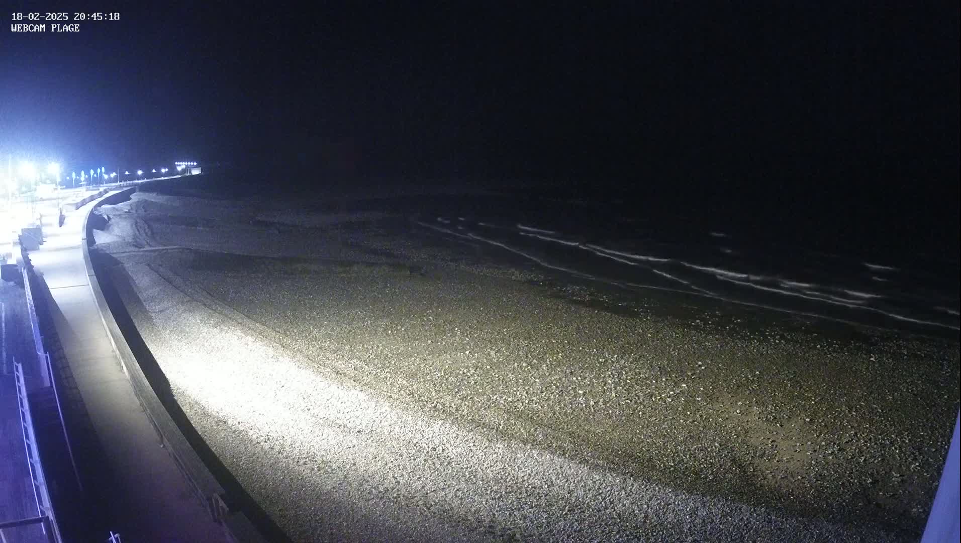 A dark, pebble beach is illuminated at night by artificial lights, with gentle waves visible in the dark water.
