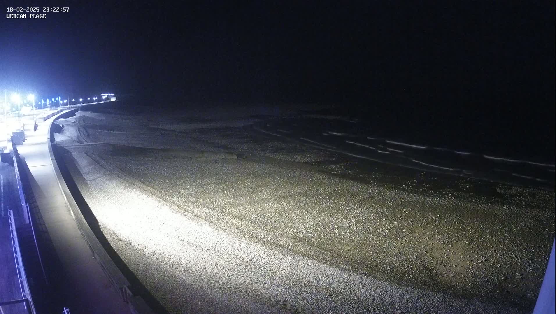 A pebble beach at night is illuminated by lights along a paved walkway bordering a dark ocean with gentle waves.