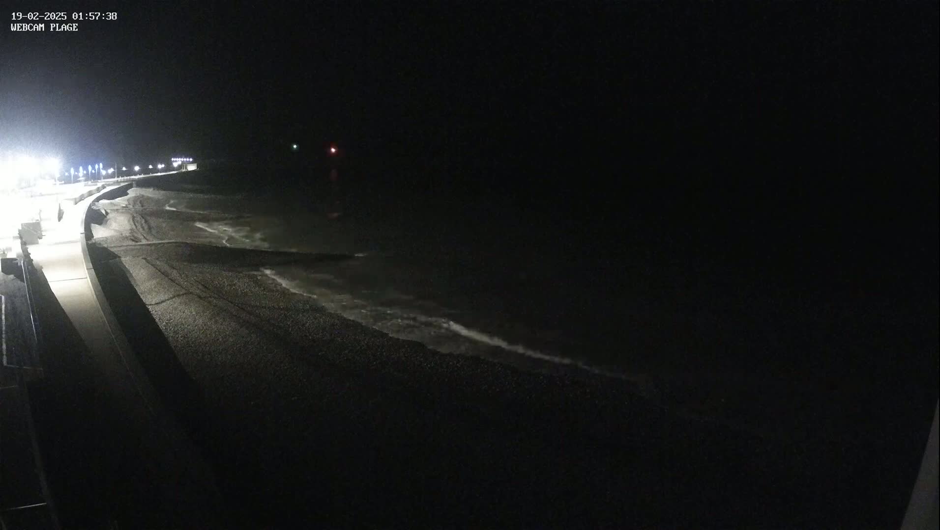 A dark, nighttime beach scene shows gentle waves lapping at a pebble shore next to a dimly lit walkway.