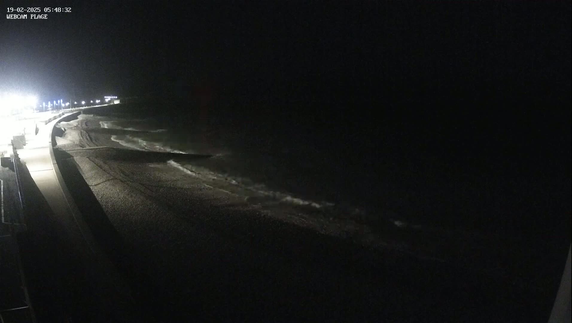 A dark, nighttime view shows a pebbled beach with gentle waves lapping the shore, illuminated by artificial lights along a paved walkway.