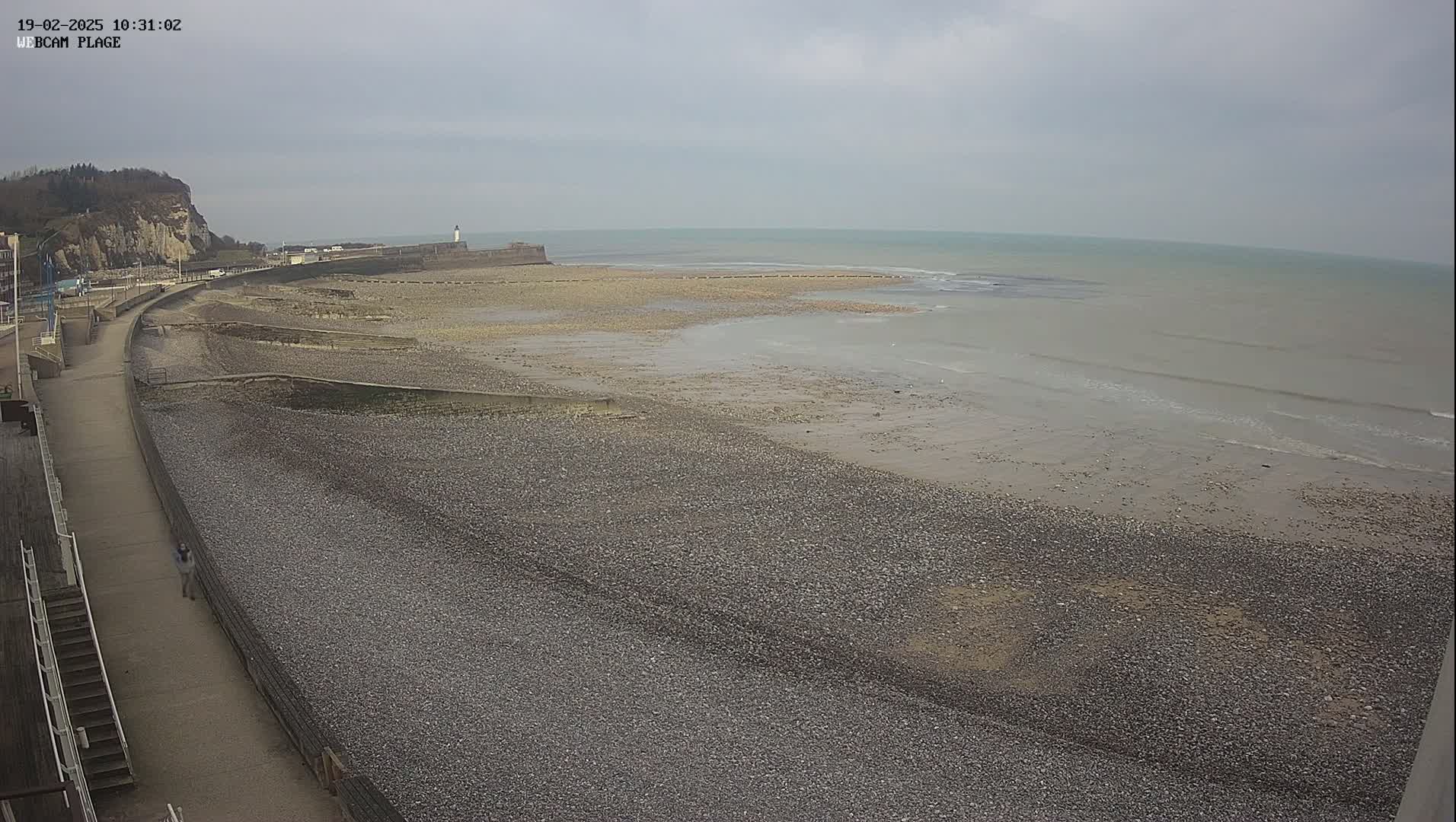 A pebble beach stretches to a calm sea under an overcast sky, with a walkway and cliffs visible in the background.