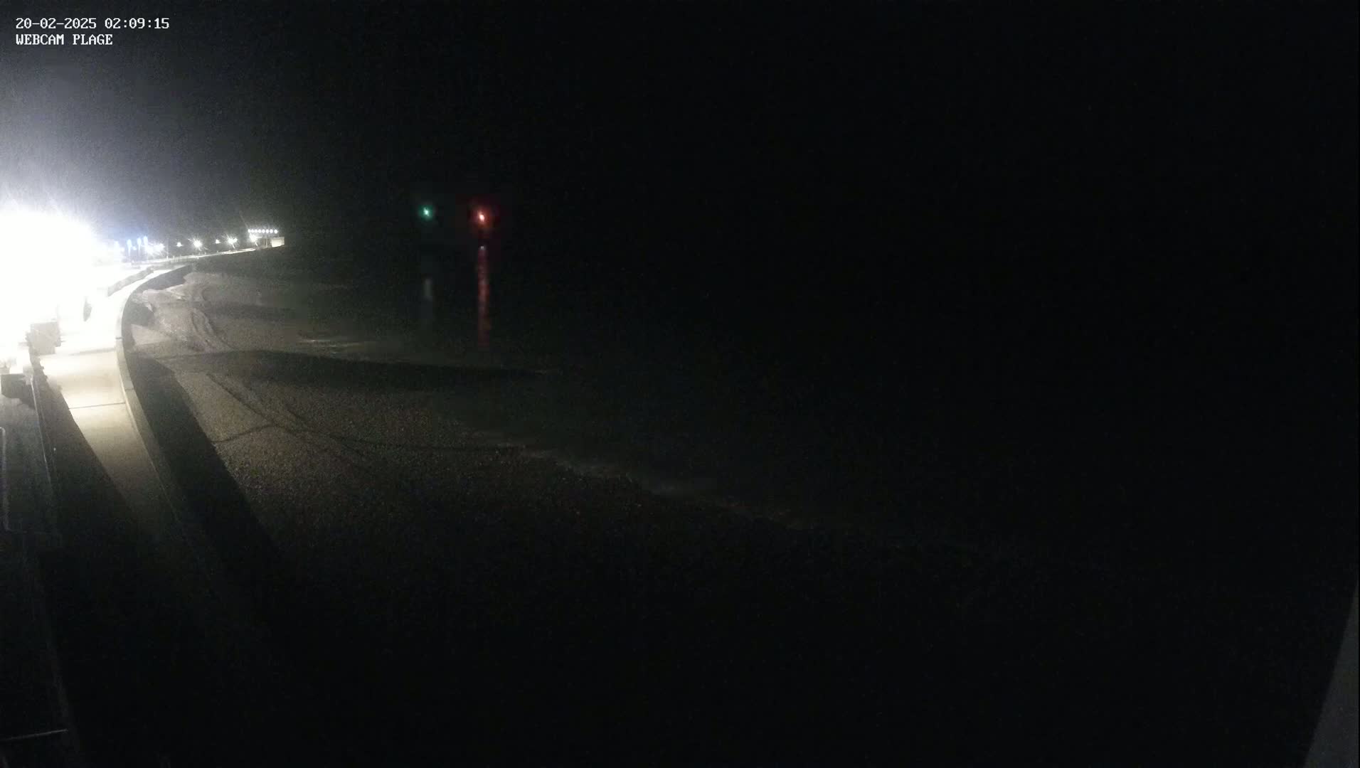 A dark, nighttime view of a pebble beach and a paved walkway, with faintly visible waves and distant lights.