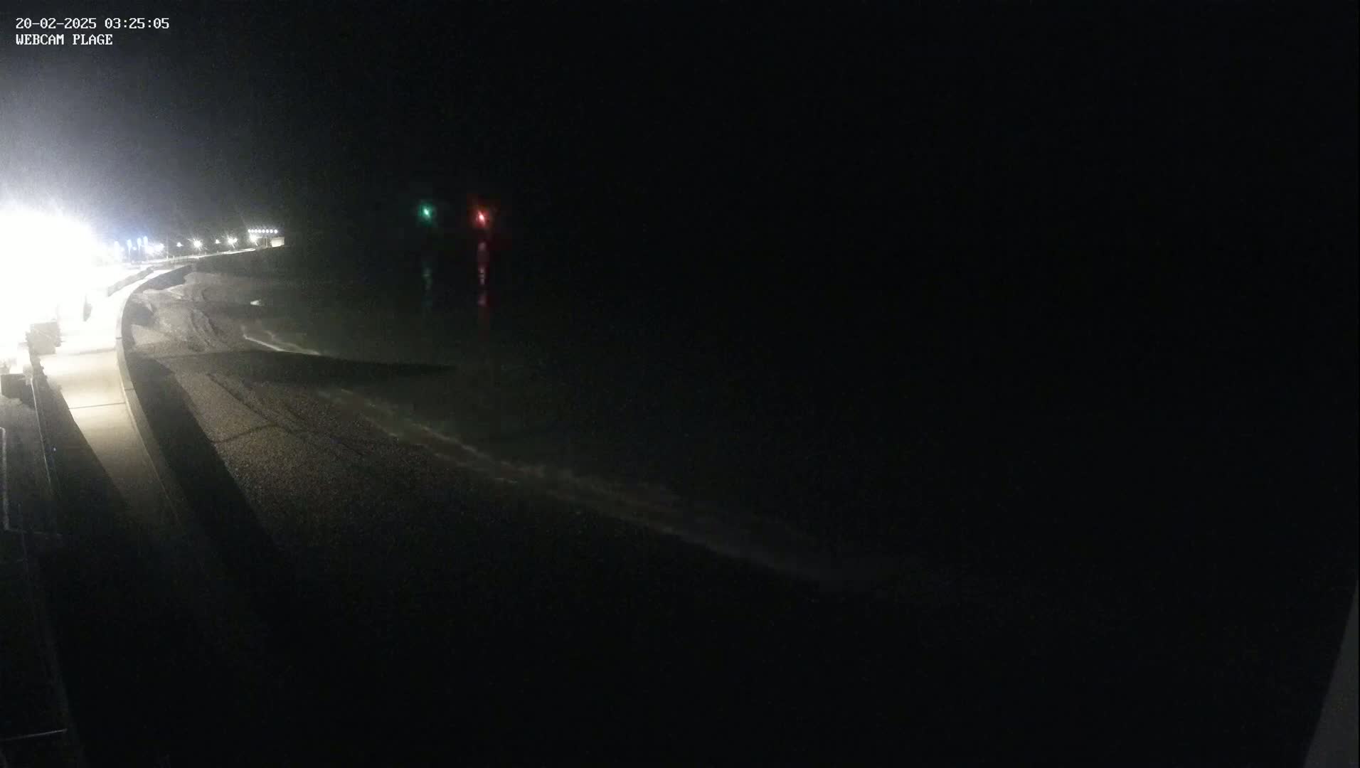 A dark, nighttime view of a pebble beach and a paved walkway with gentle waves lapping the shore.