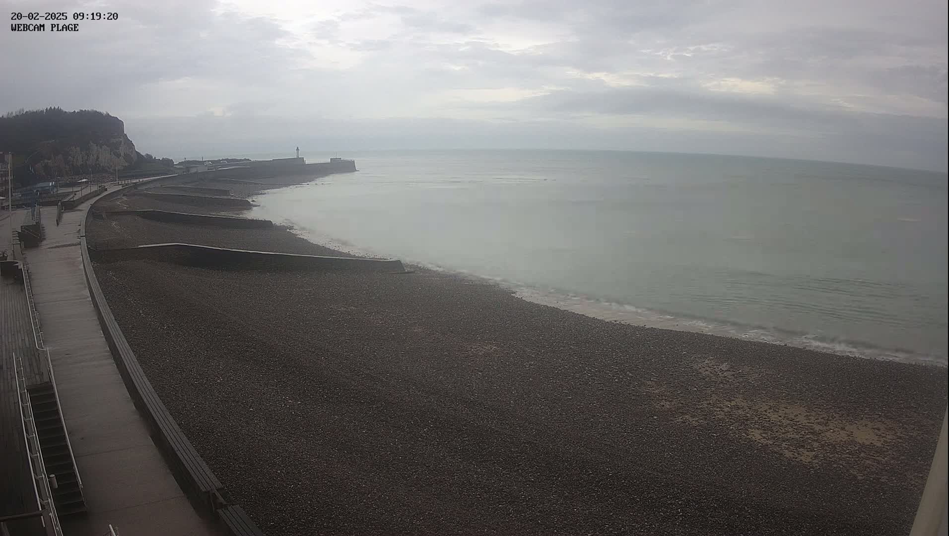 A pebble beach stretches along a sea wall under an overcast sky, with calm waves lapping the shore.