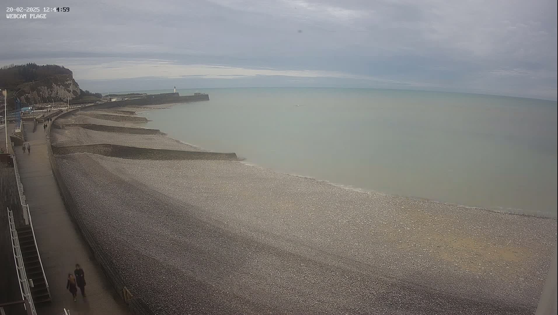 A pebble beach stretches along a coastline under an overcast sky, with a few people walking along a paved path above the shore.