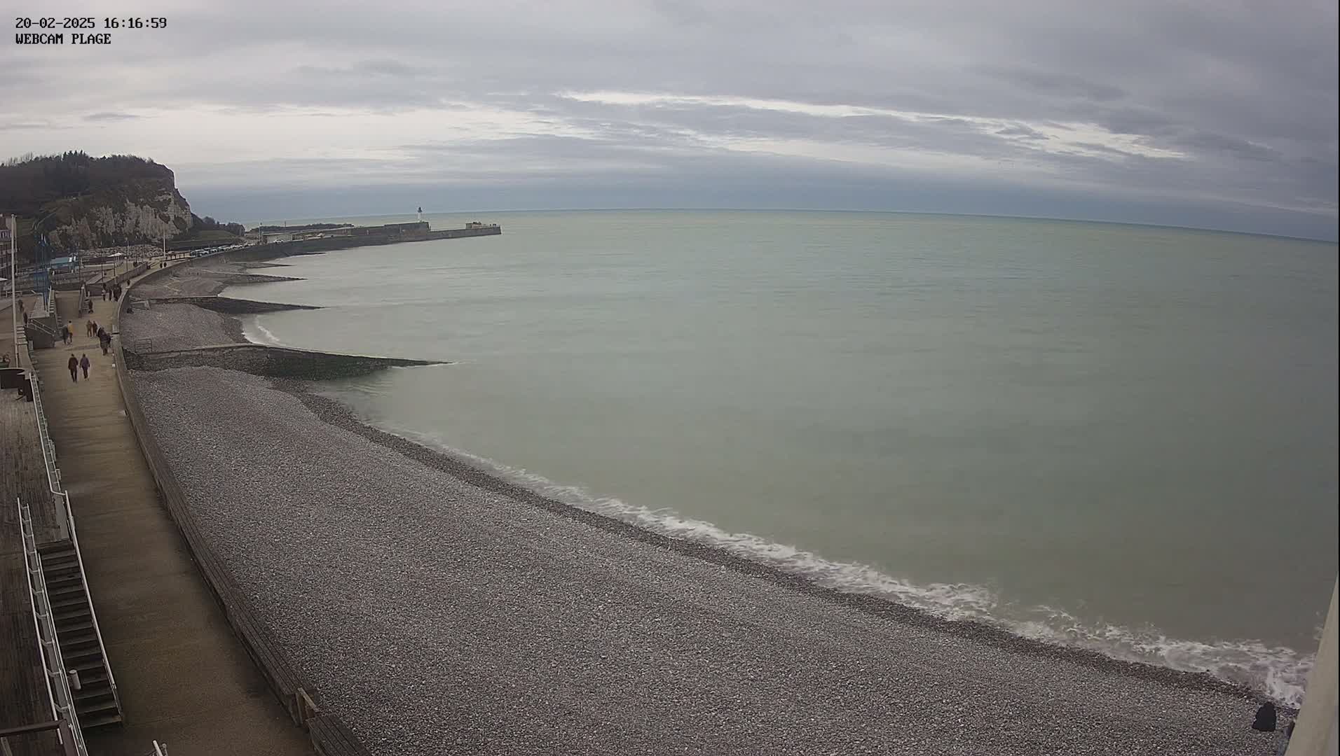 A pebble beach stretches along a calm, grey sea under an overcast sky, with a walkway and people visible in the distance.